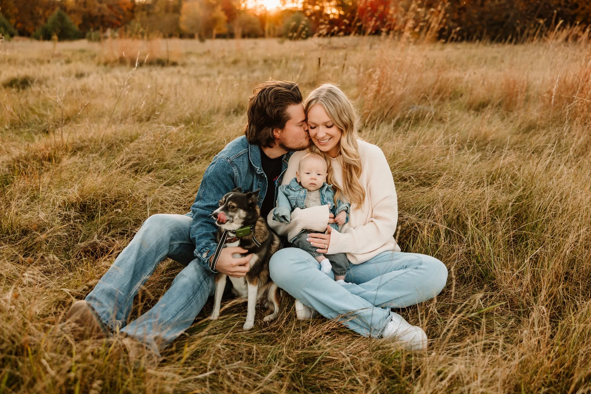 A happy family of three with a dog sitting in a grassy field during sunset. The man is kissing the woman on the cheek, and the woman is holding a small child. The dog, a husky mix, is sitting on the woman's lap.