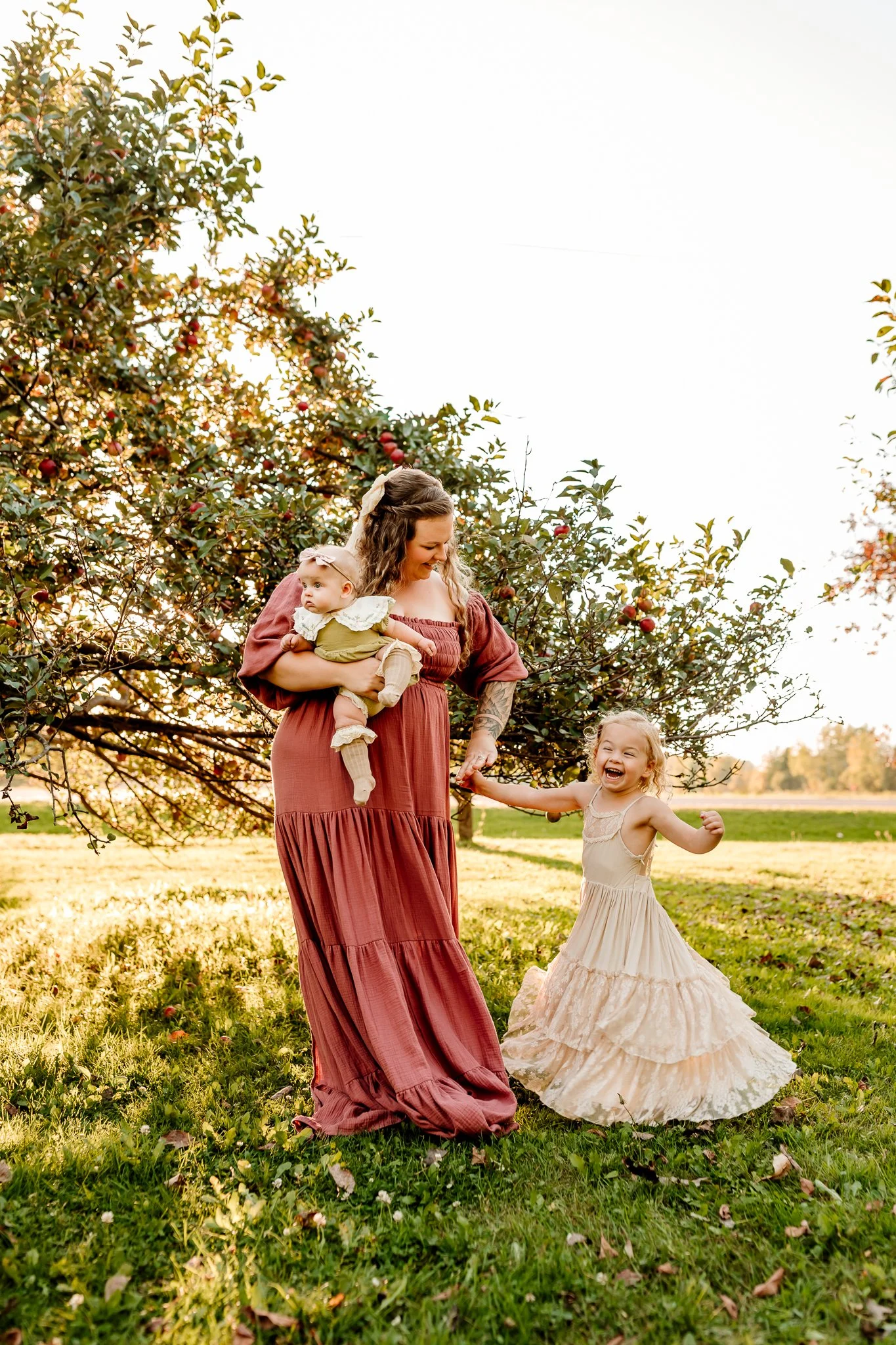 A woman with two young children outdoors in a grassy area with trees. The woman holds a baby, and the older girl is dancing and smiling. The scene is bathed in warm, natural light.