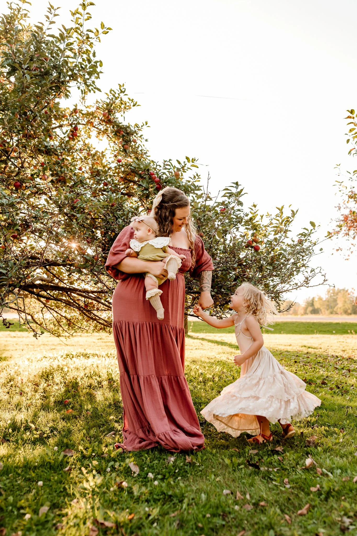 A woman with two young children under an apple tree in a grassy field during sunset.