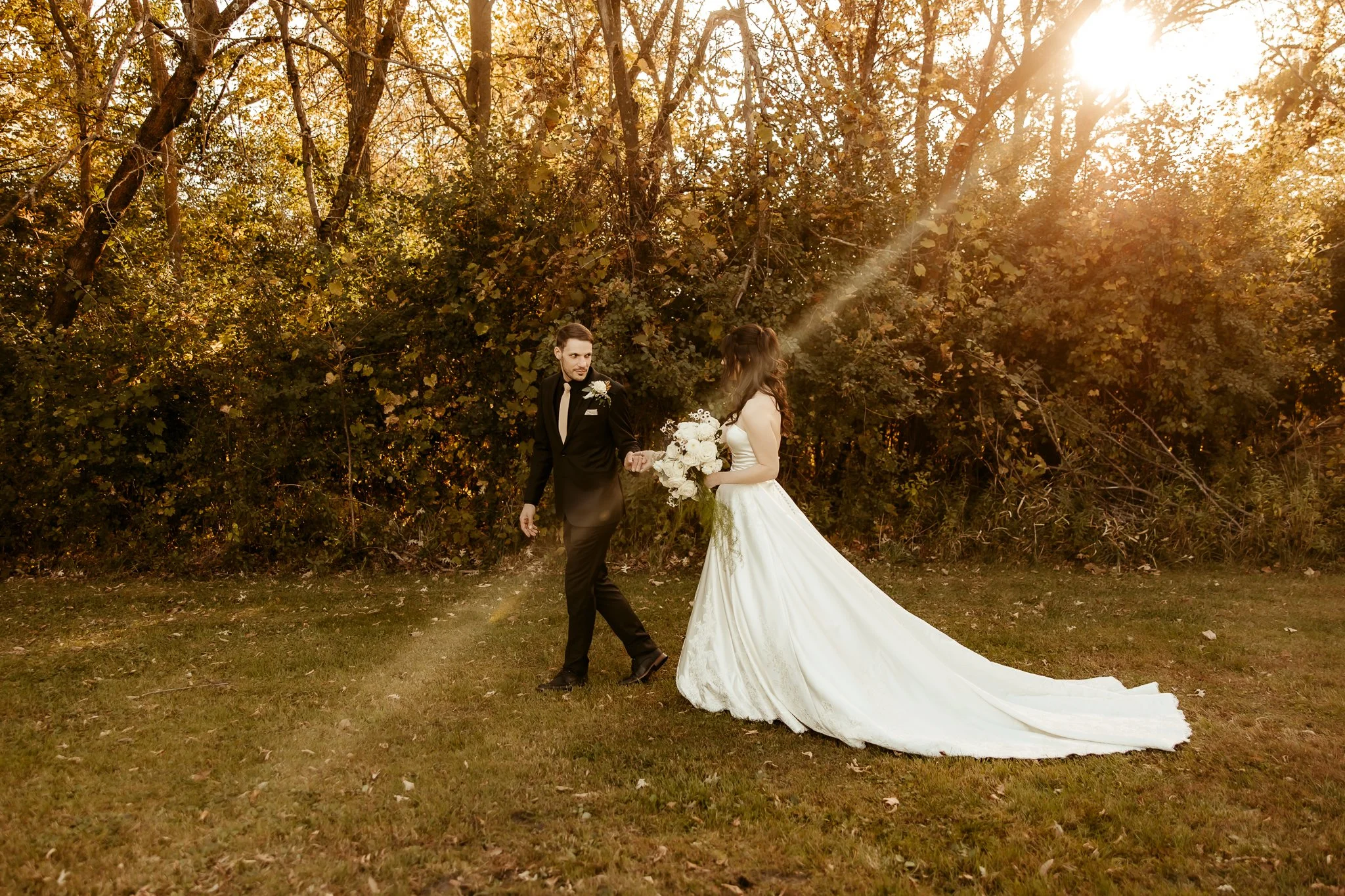 A bride and groom holding hands in a park at sunset. The bride wears a long white wedding gown and holding a bouquet, while the groom is dressed in a black tuxedo. Sunlight filters through trees in the backgroundPine city minnesota wedding photograph
