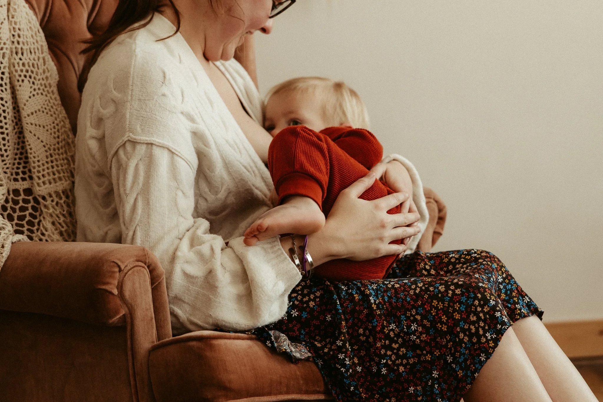 A woman sits on a couch holding a young child in her arms, breastfeeding. The woman wears a cream-colored sweater with a cable-knit pattern, and the child wears a red outfit. Another person is sitting beside her, partially visible.
