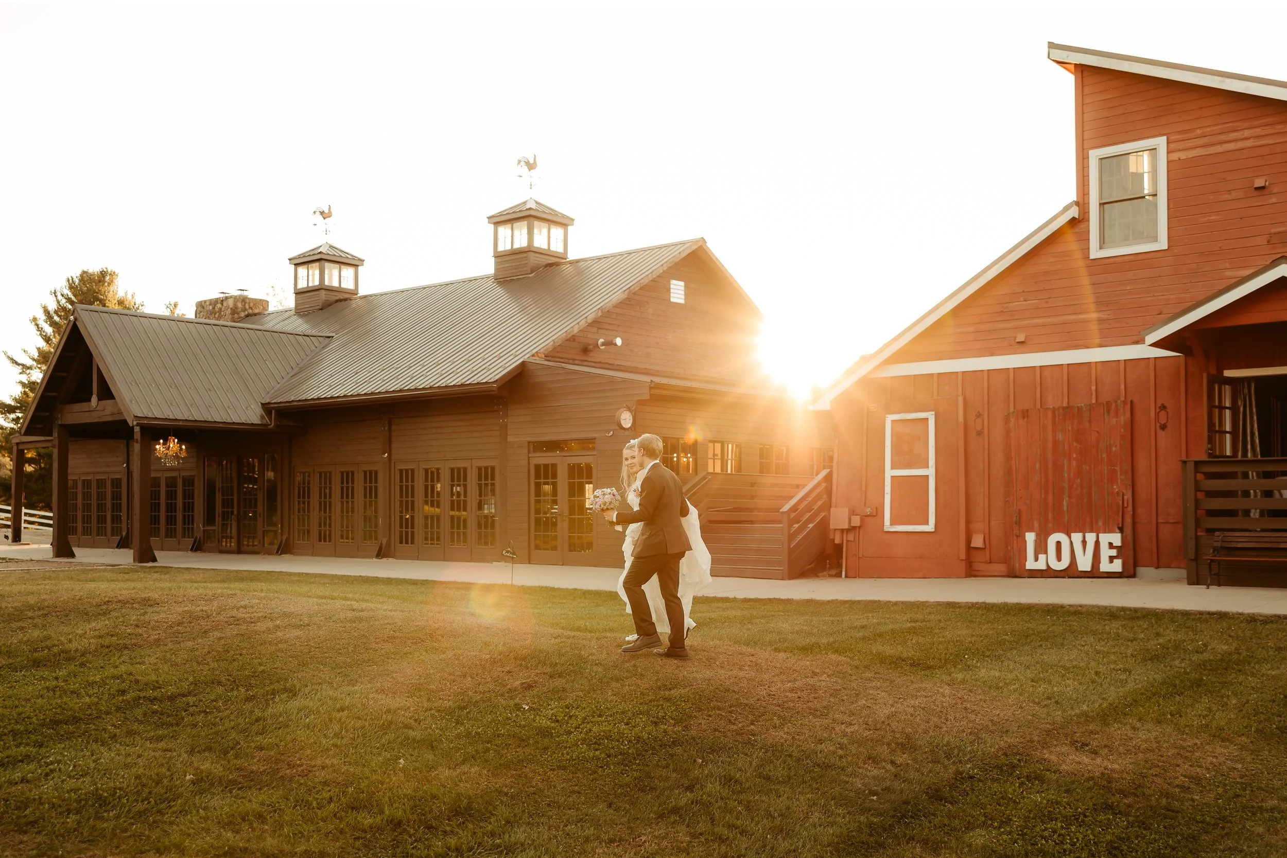 A couple walking on grass holding hands at sunset, with a barn-style building in the background that has a sign reading 'LOVE' on the red barn wall.