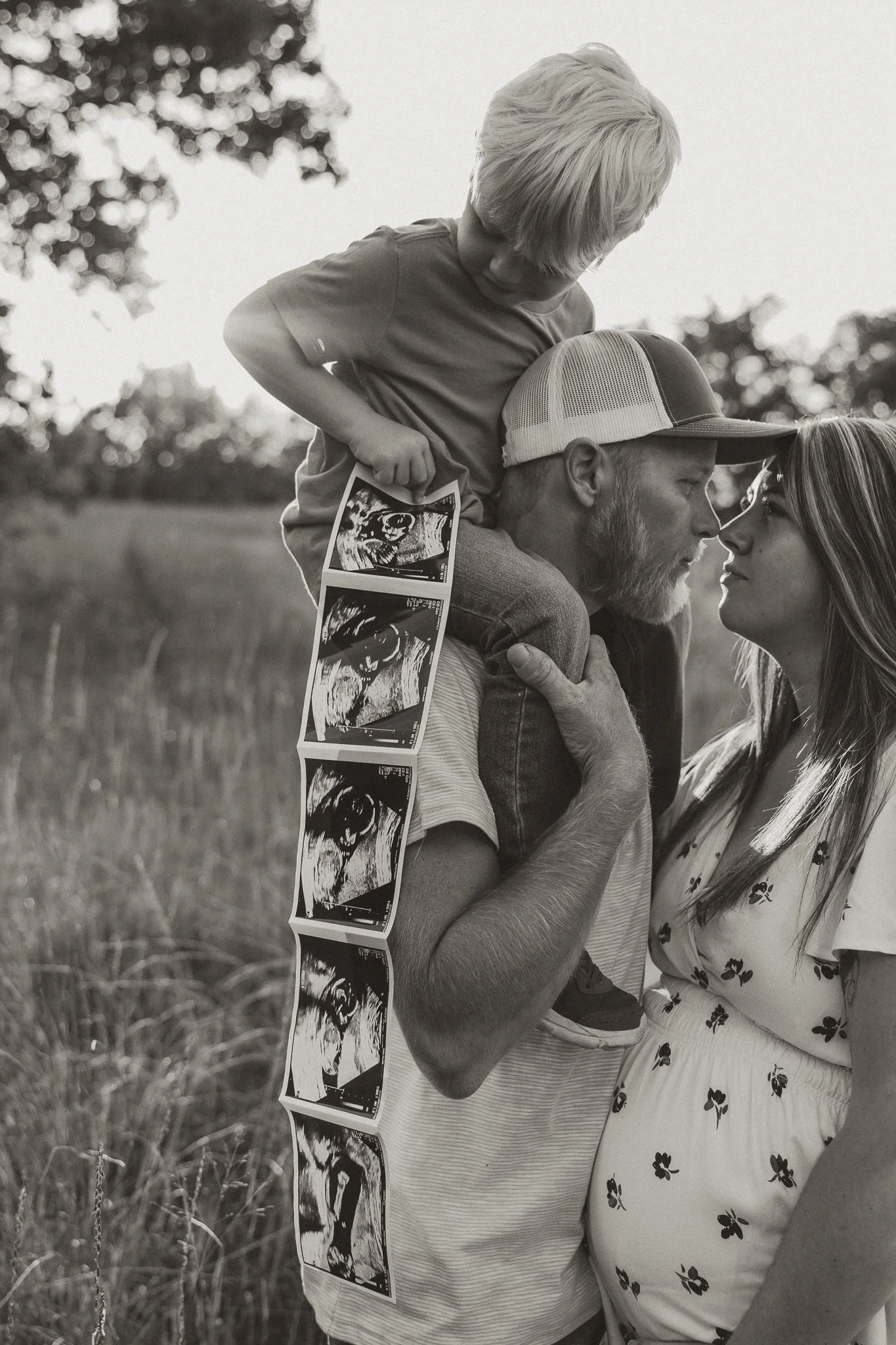 A couple and a child outdoors in a field, with the man carrying ultrasound images on his arm, while the woman and the child look at each other