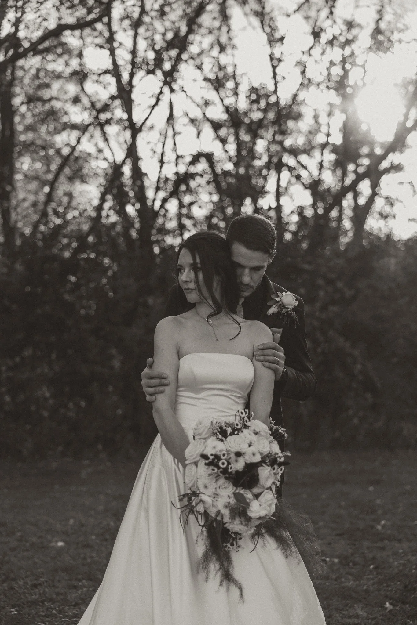 Black and white photo of a bride holding a bouquet with a groom standing behind her, both outdoors surrounded by trees at sunset.Pine city minnesota wedding photography 