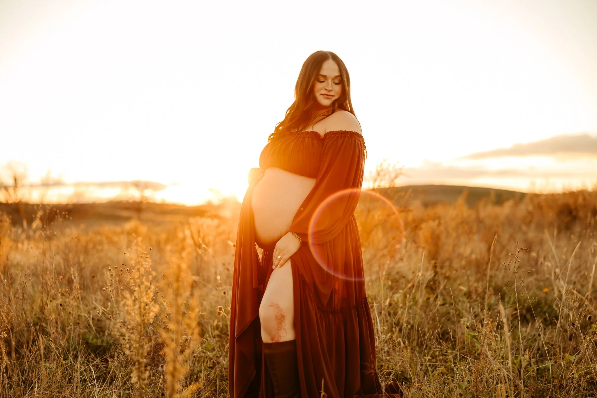 A pregnant woman in a flowing dark dress stands in a field at sunset, gently holding her belly and looking down with a peaceful expression.