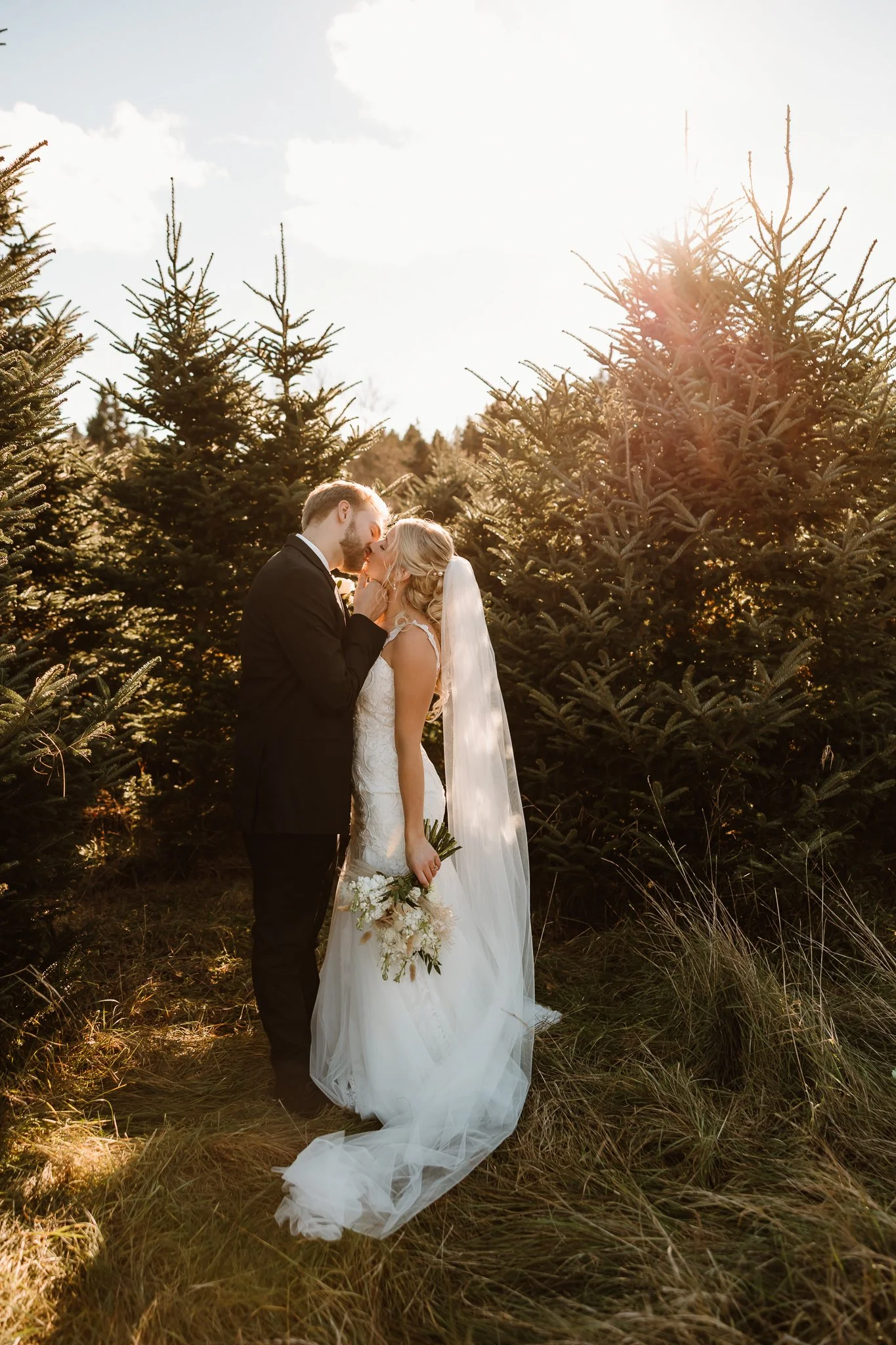A bride and groom kiss in a field of pine trees during sunset, with the bride holding a bouquet and wearing a white wedding gown with a long veil, and the groom in a black suit.