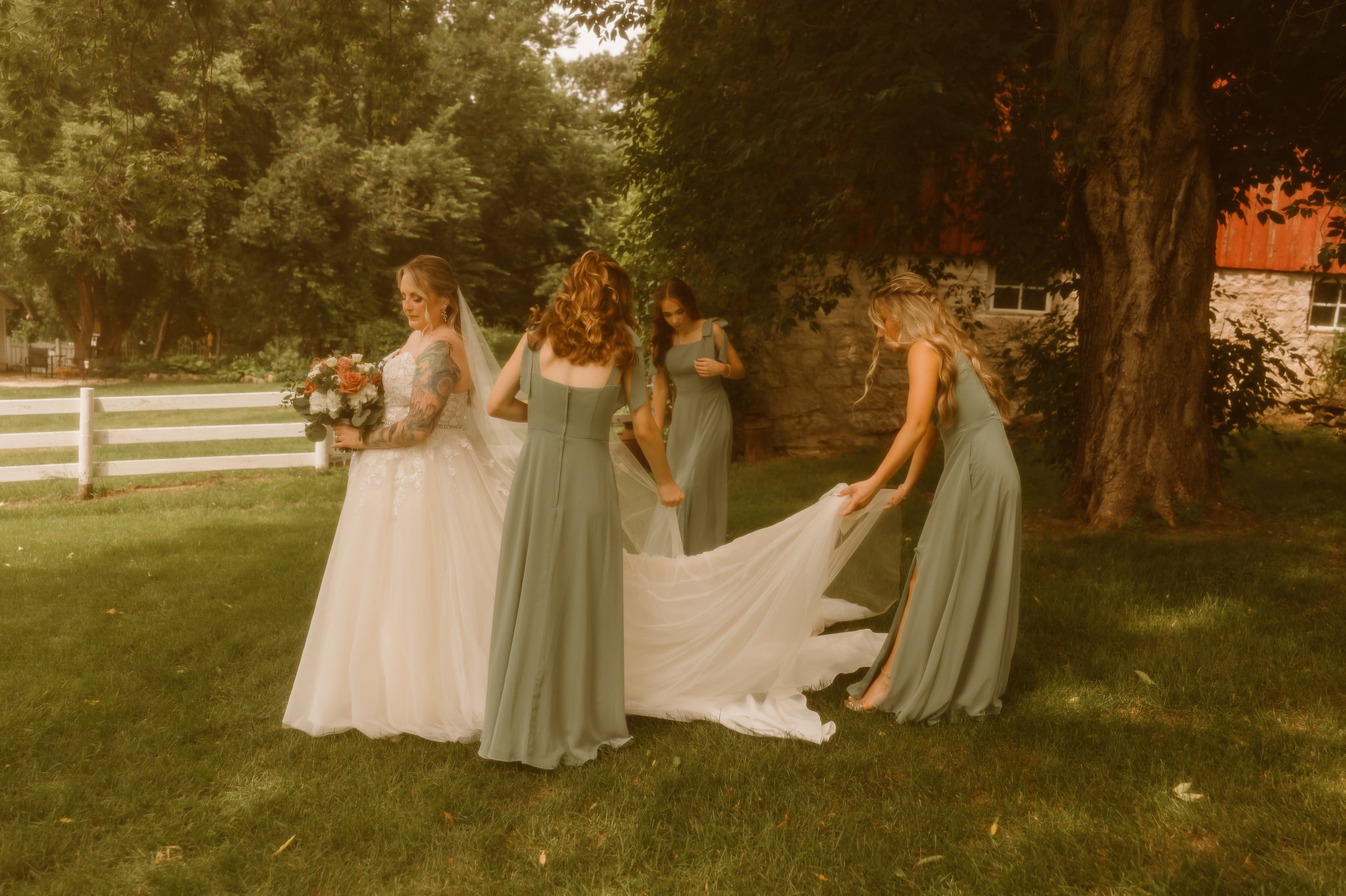 Bride in white wedding dress with bouquet, four bridesmaids in green dresses arranging her train outdoors under a large tree with a rustic building in the background.
