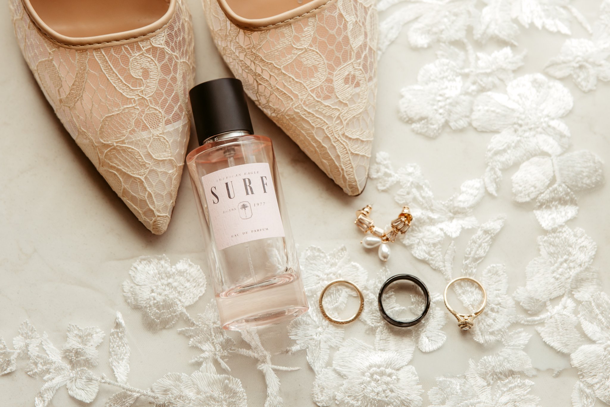 A pair of cream-colored lace high heels, a bottle of perfume labeled 'Surf,' three rings (one gold, one black, one pearl with a gold band), and a pair of pearl earrings on a white embroidered fabric surface.