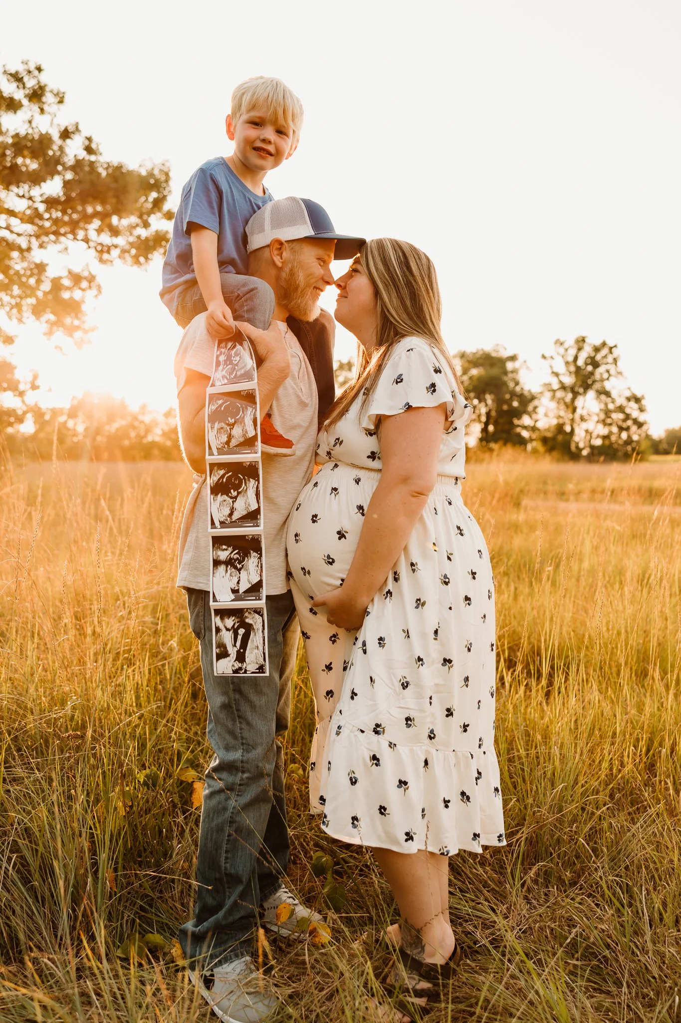 A pregnant woman and a man with a child on his shoulders are in a field at sunset, sharing a close moment.