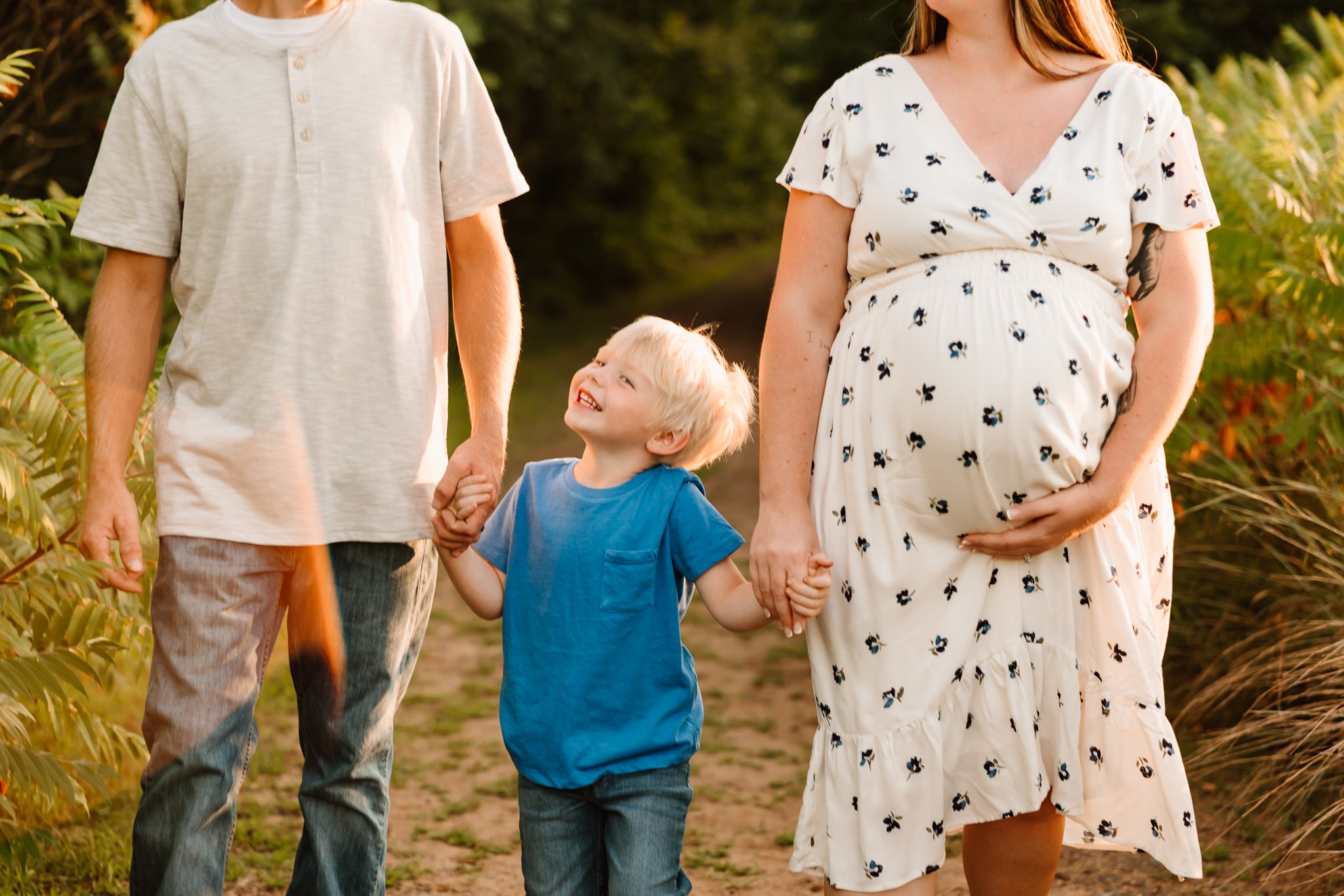 A pregnant woman and her partner holding hands with a young boy while walking outdoors on a sunny day.