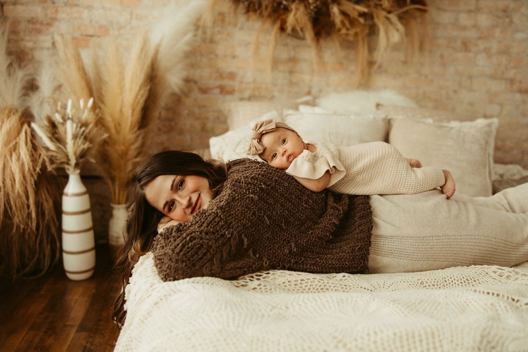 A woman lying on a bed with a young child on her back, both smiling. The woman has dark hair and is wearing a brown knitted sweater. The child has a bow headband and light-colored clothing. The room has a cozy, rustic style with beige pillows, a bric