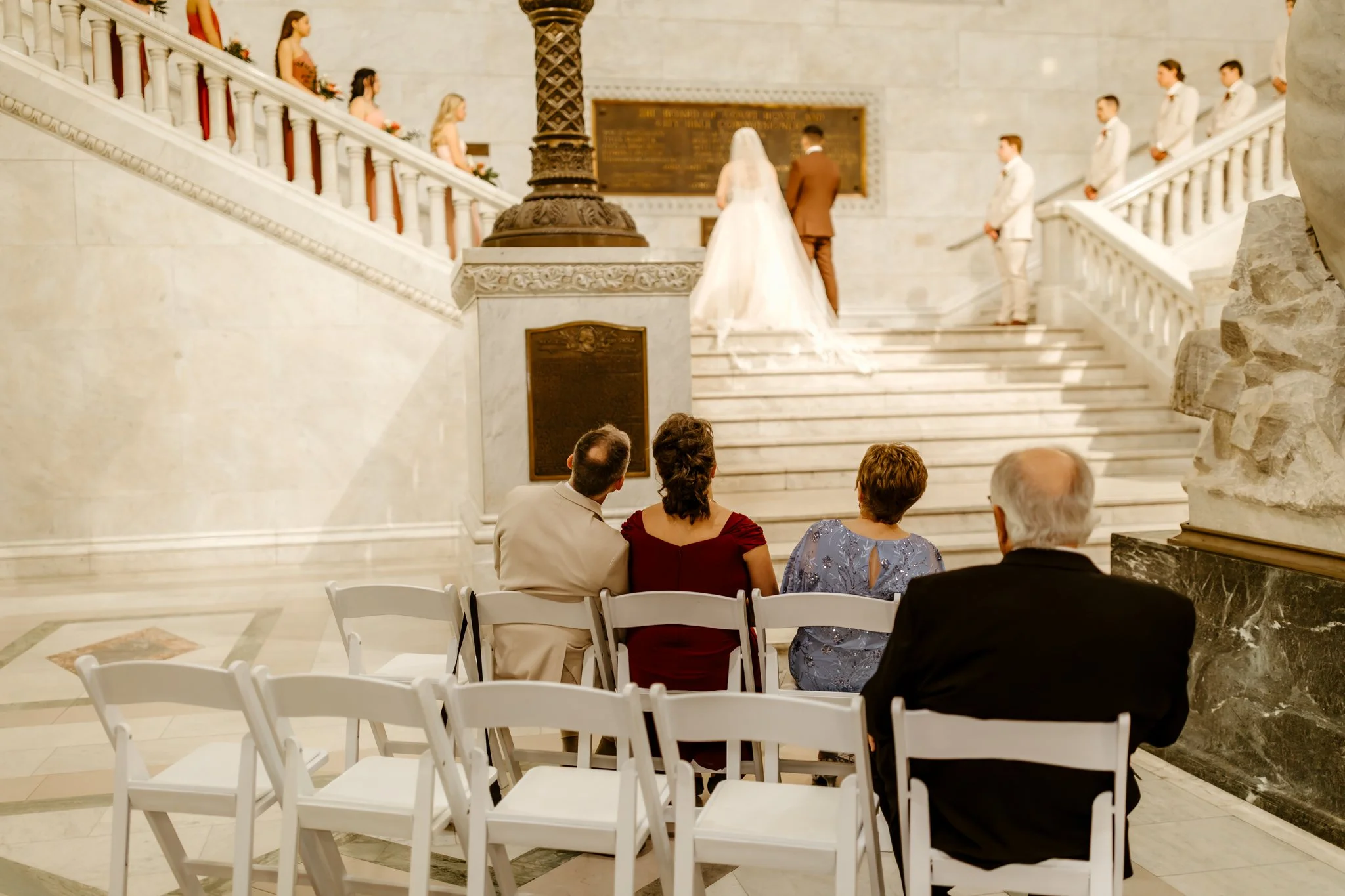 People seated in white chairs watching a wedding ceremony in a grand marble hall with a staircase and a bride and groom at the altar.