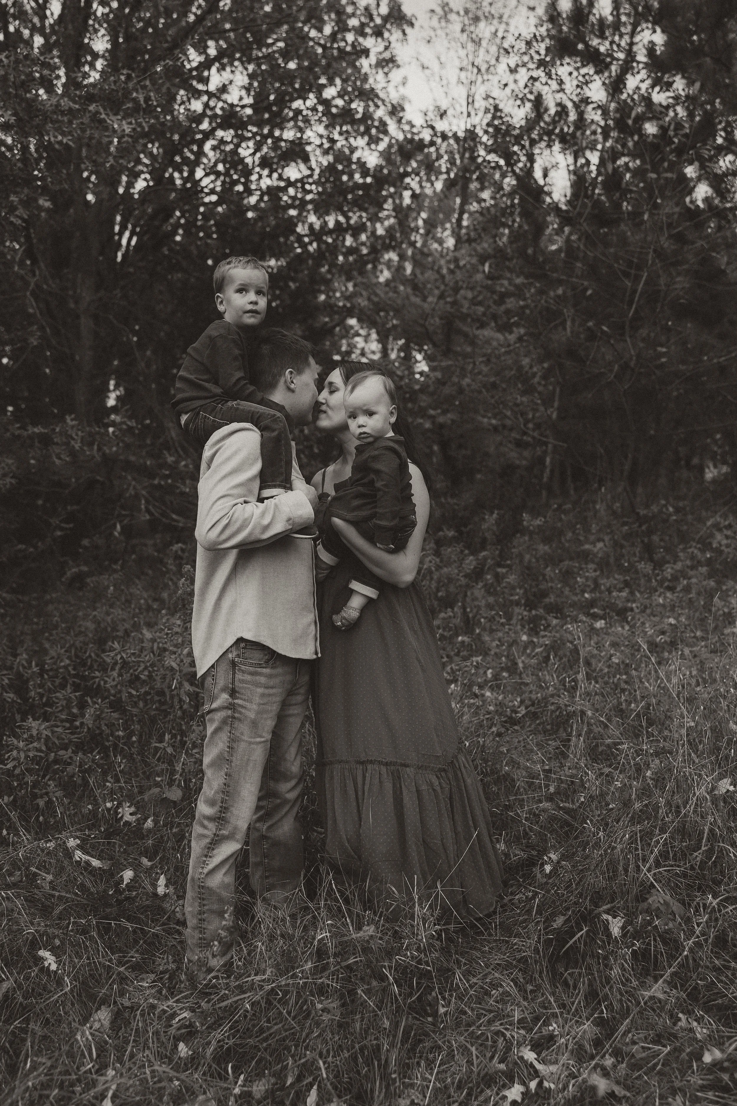 A family of four standing outdoors in a wooded area, with trees and foliage in the background. The father is holding a young child on his shoulder, the mother is holding a baby, and all are facing the camera. The image is in black and whiteMinnesota 
