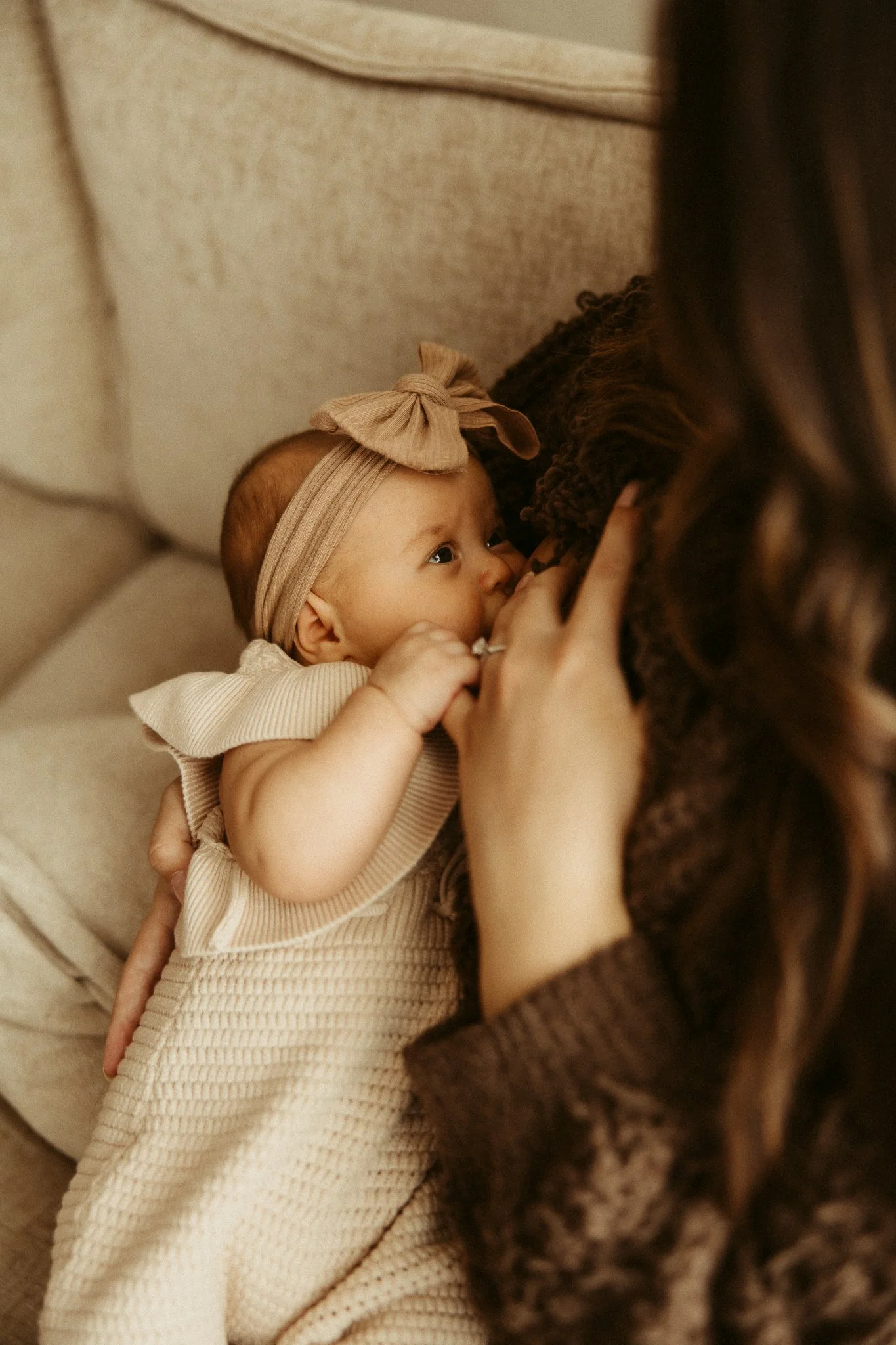 A baby girl with a bow headband breastfeeding while lying on a beige sofa with her mother, who has long brown hair.