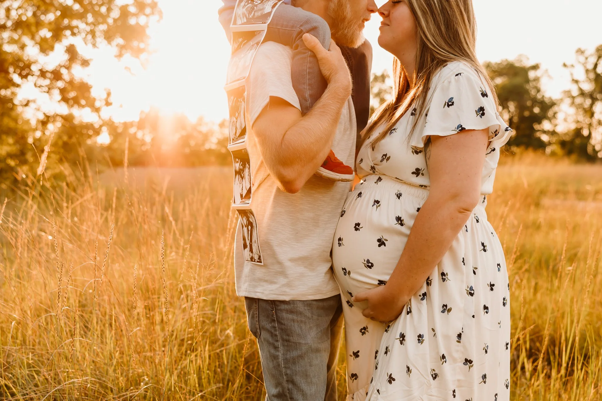 A pregnant woman and a man standing close together in a field at sunset, face to face, with the man's arms around the woman's shoulders and her hand on her belly.
