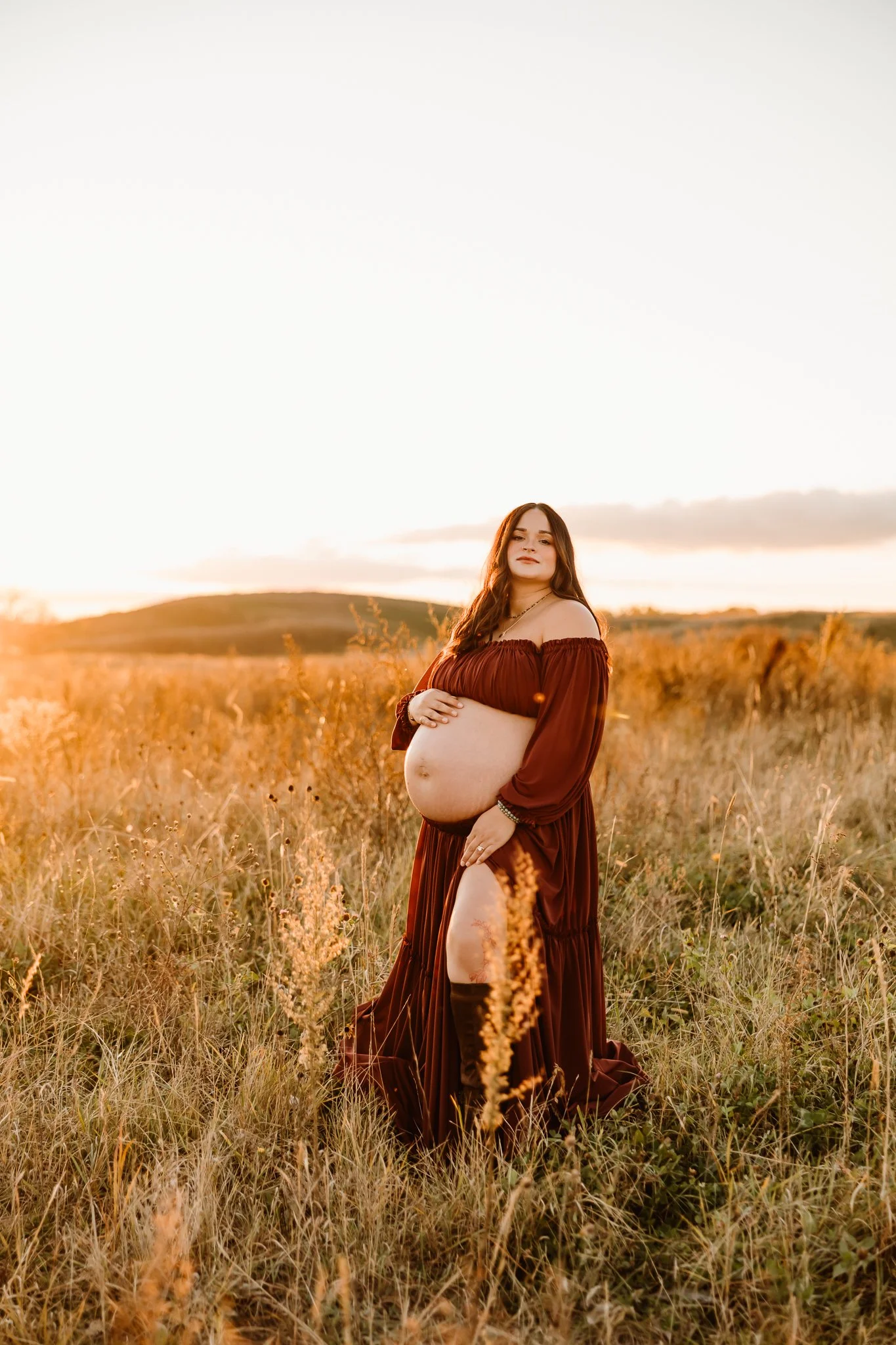 Pregnant woman in a long brown dress standing in a grassy field during sunset.