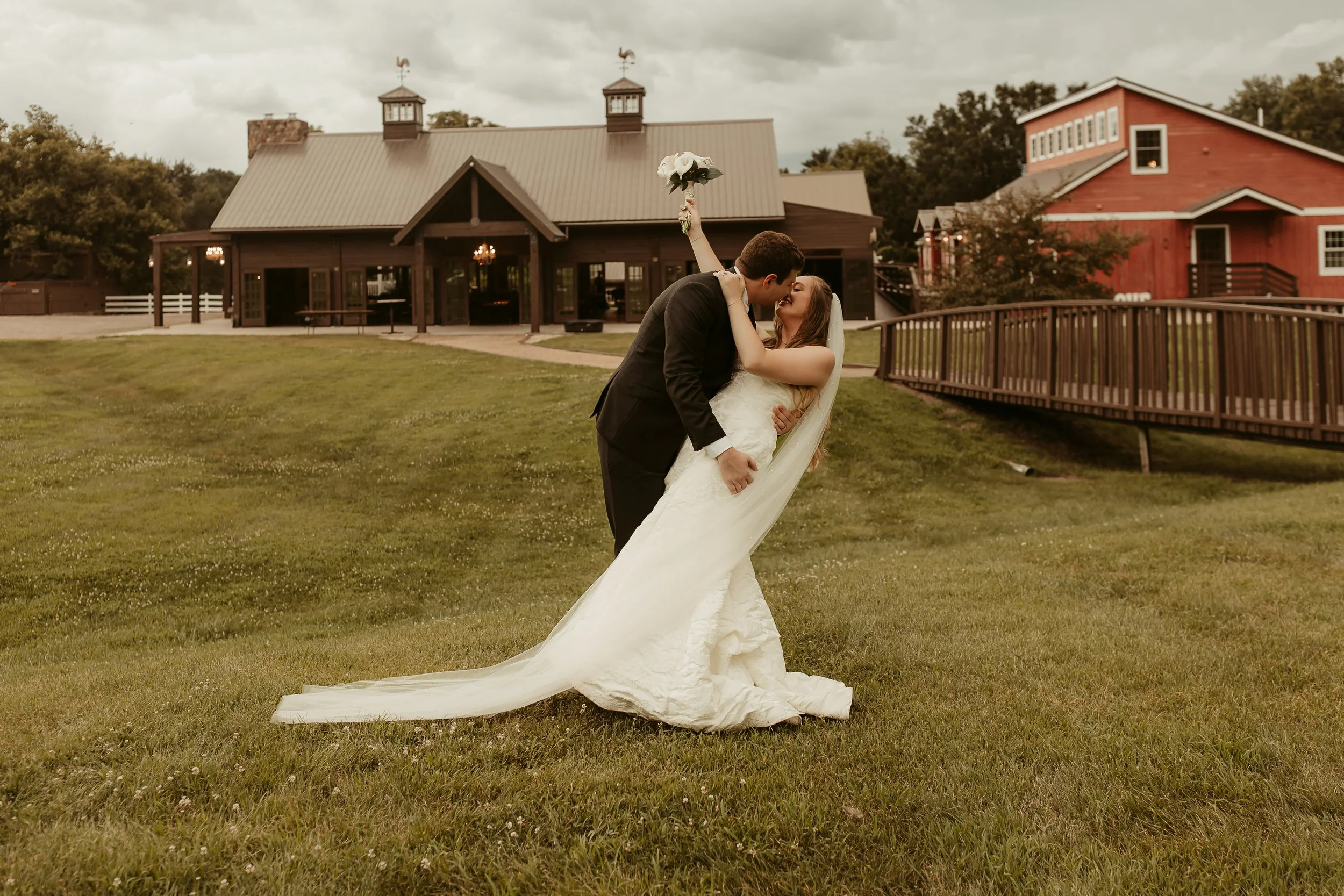 A bride and groom sharing a kiss outside on a lawn, with the groom in a black suit and the bride in a white wedding dress holding a bouquet of white flowers above their heads, with buildings and a cloudy sky in the background.