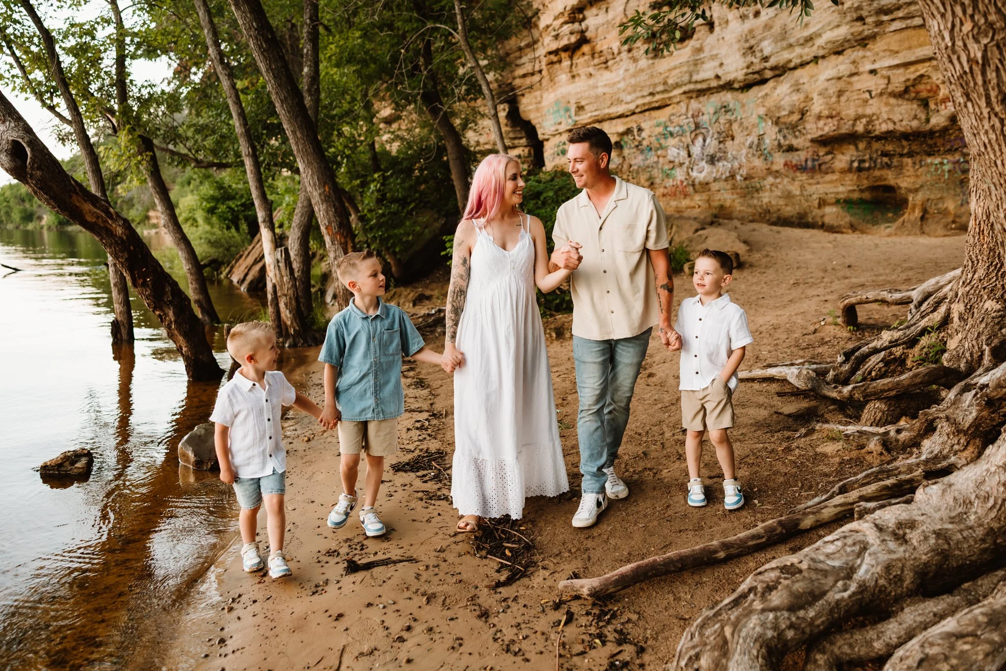 A family of six walking along a sandy riverside with trees and a rocky cliff in the background, holding hands and smiling.