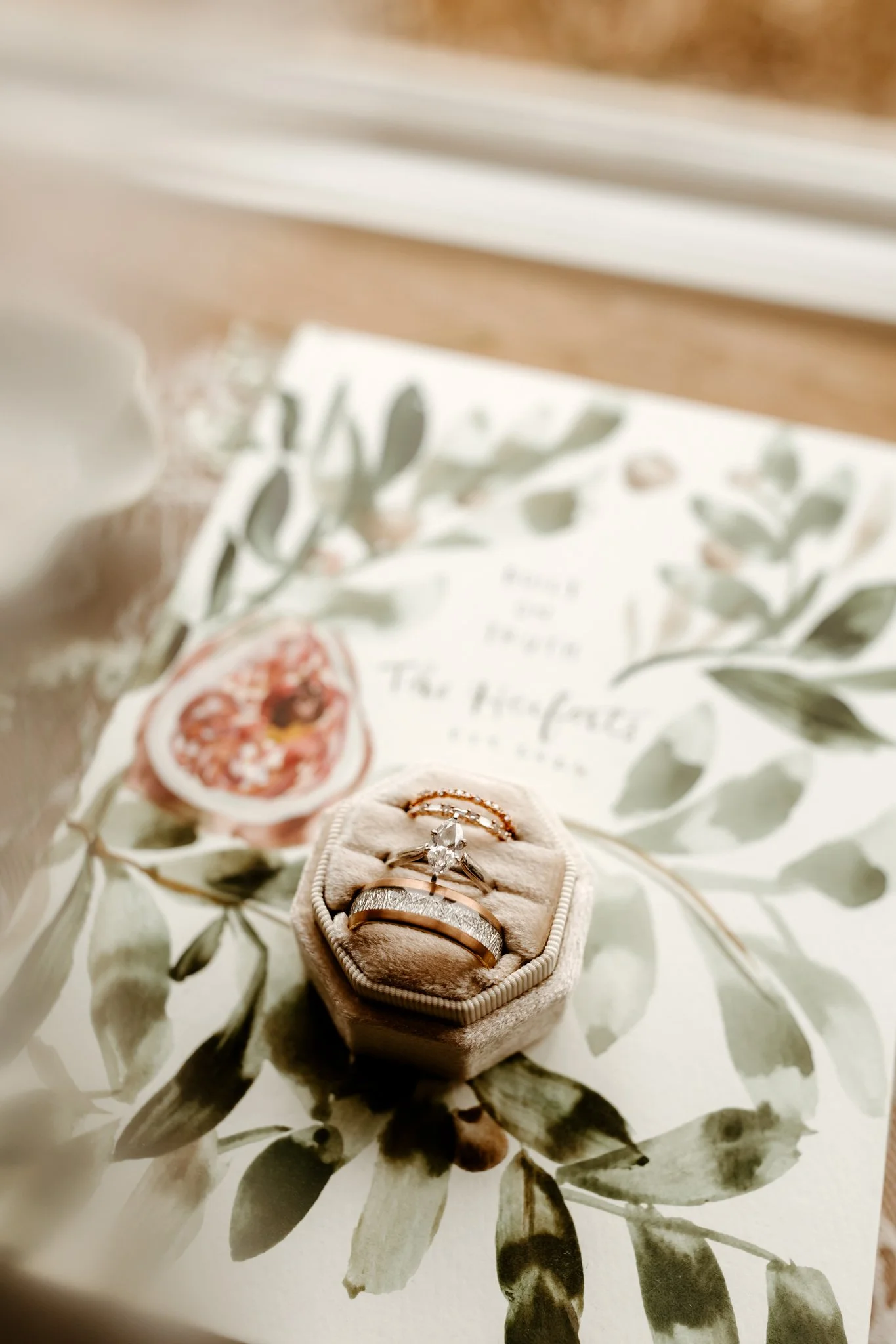 A jewelry display with a diamond engagement ring and wedding bands in a velvet ring box, placed on a floral invitation with green leaves and pink flowers.