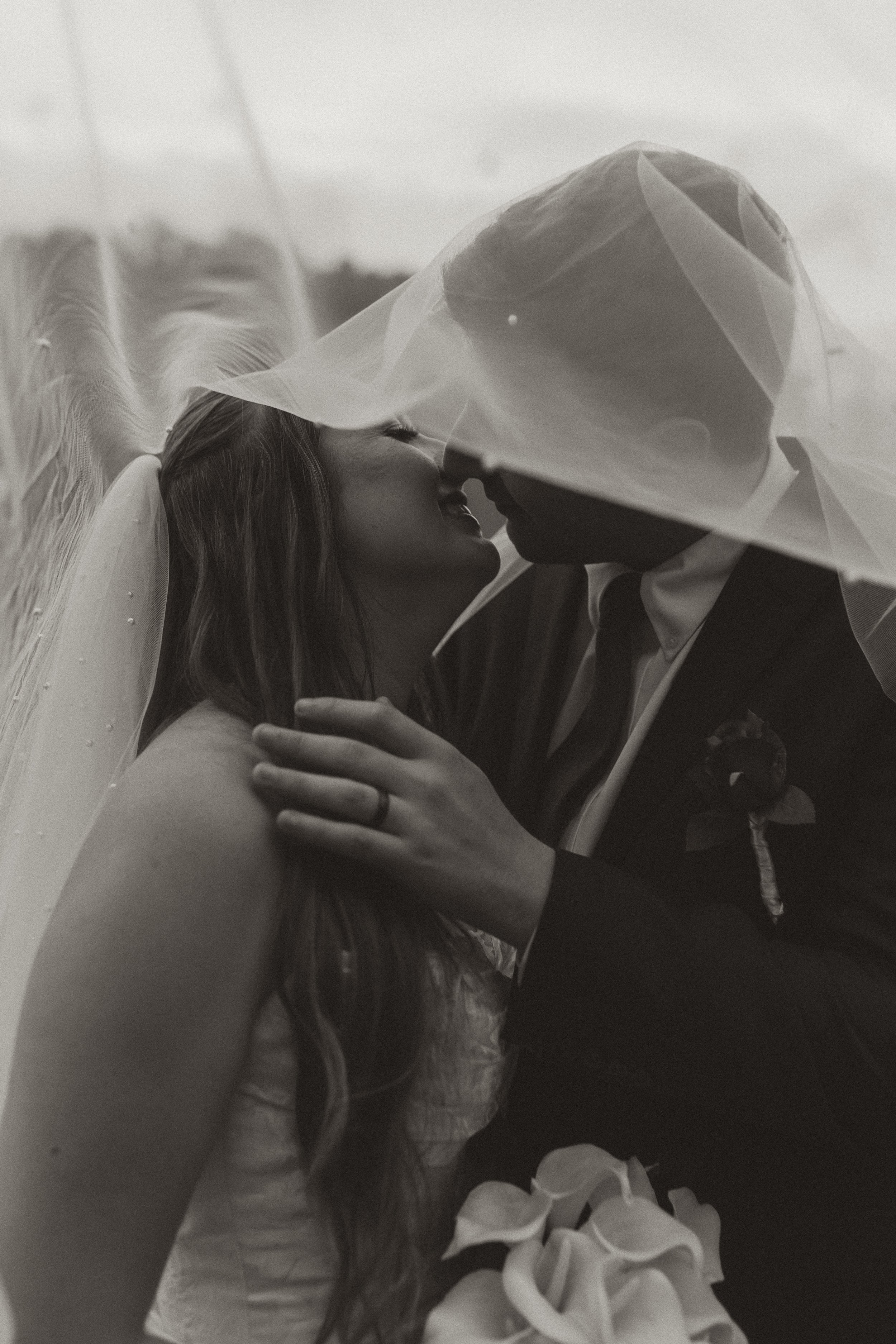 A black and white photo of a bride and groom sharing a kiss under the bride's veil, with the bride holding a bouquet of calla lilies.
