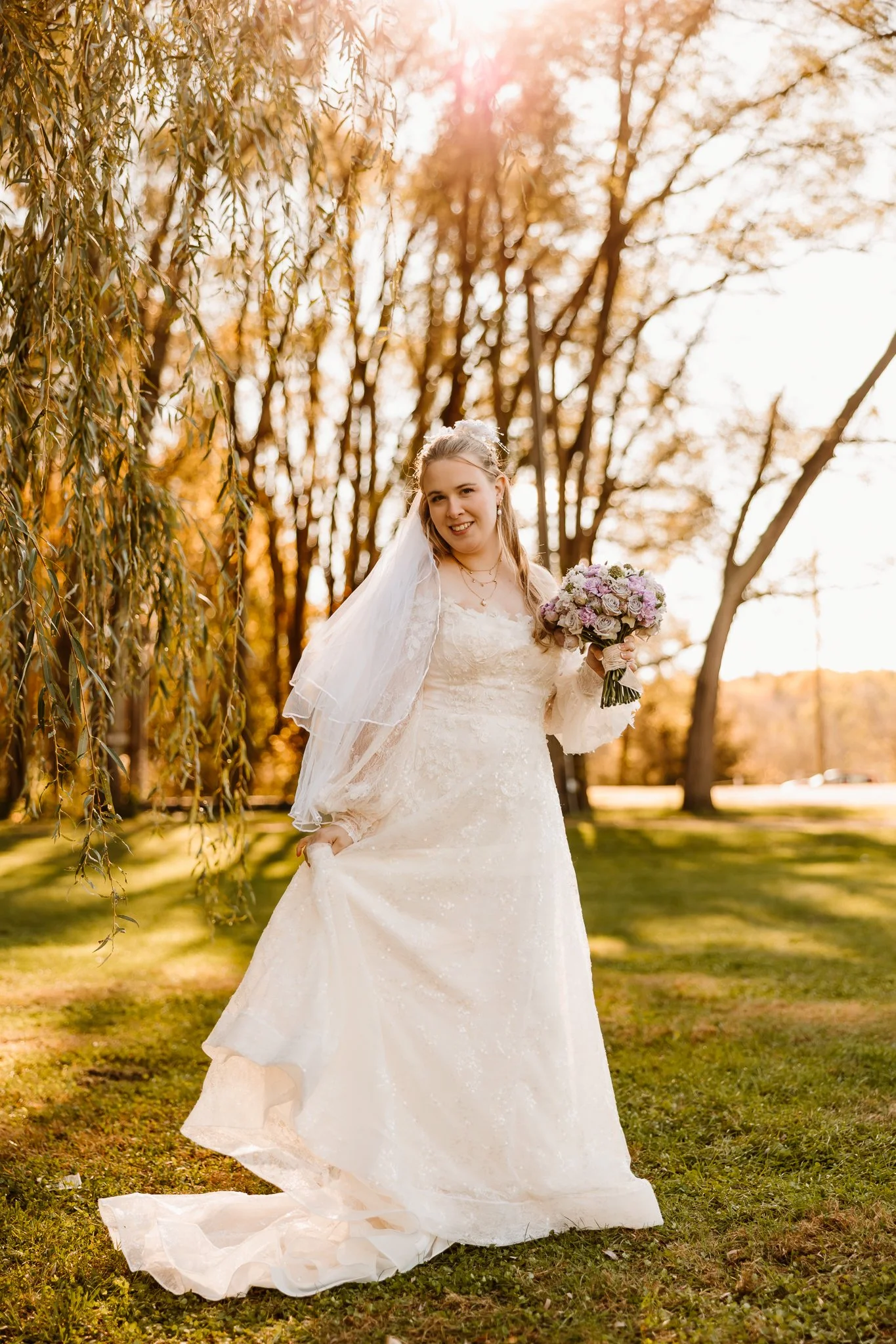 A smiling bride in a lace wedding dress holding a bouquet of flowers in an outdoor park setting during golden hour.