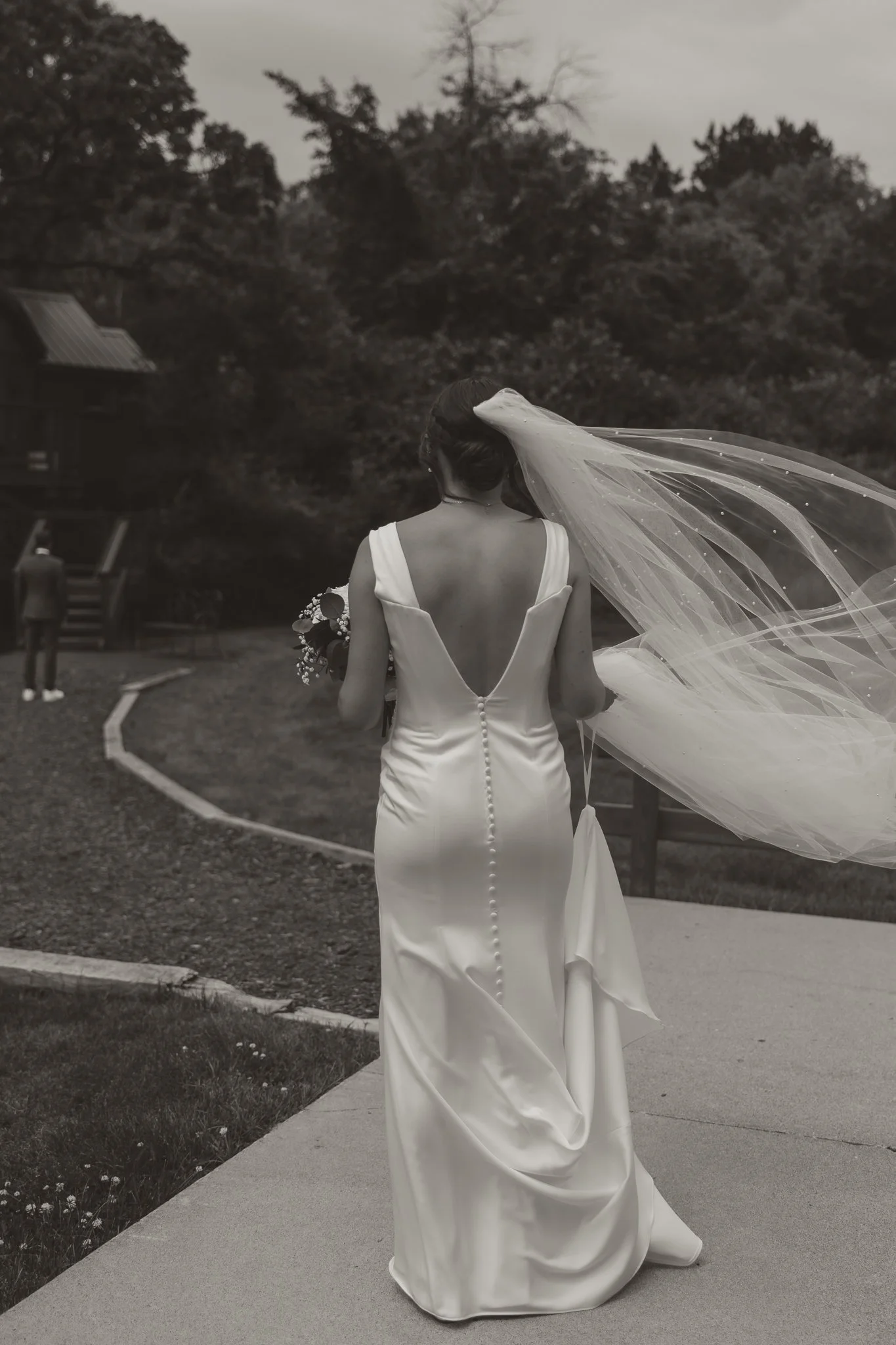 A bride in a strapless wedding dress with buttons down the back, holding a bouquet, walking outdoors with a long veil flowing behind her, and a man is visible in the distance.
