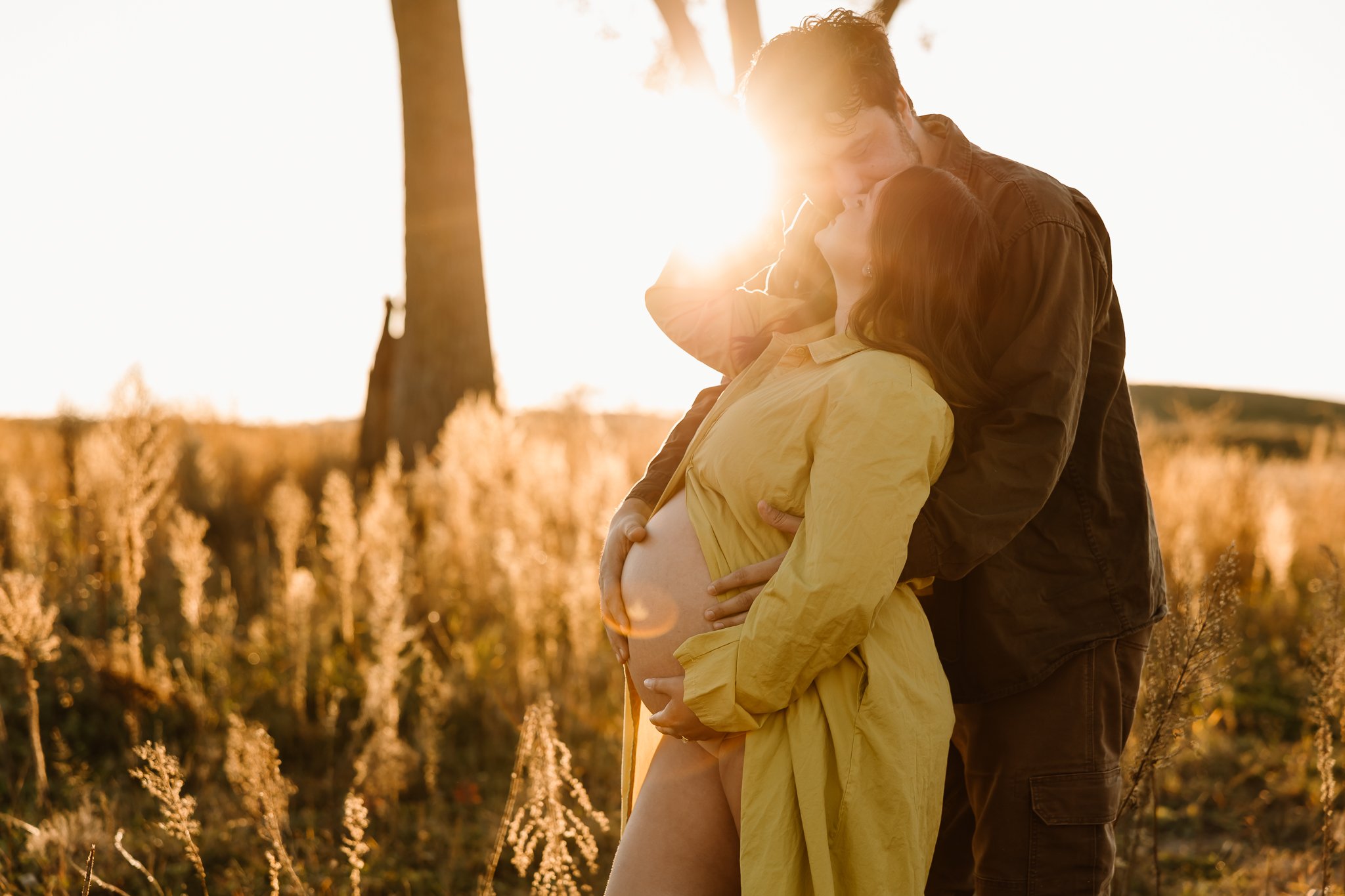 A couple embracing outdoors at sunset, with the woman pregnant and the sun shining behind them.
