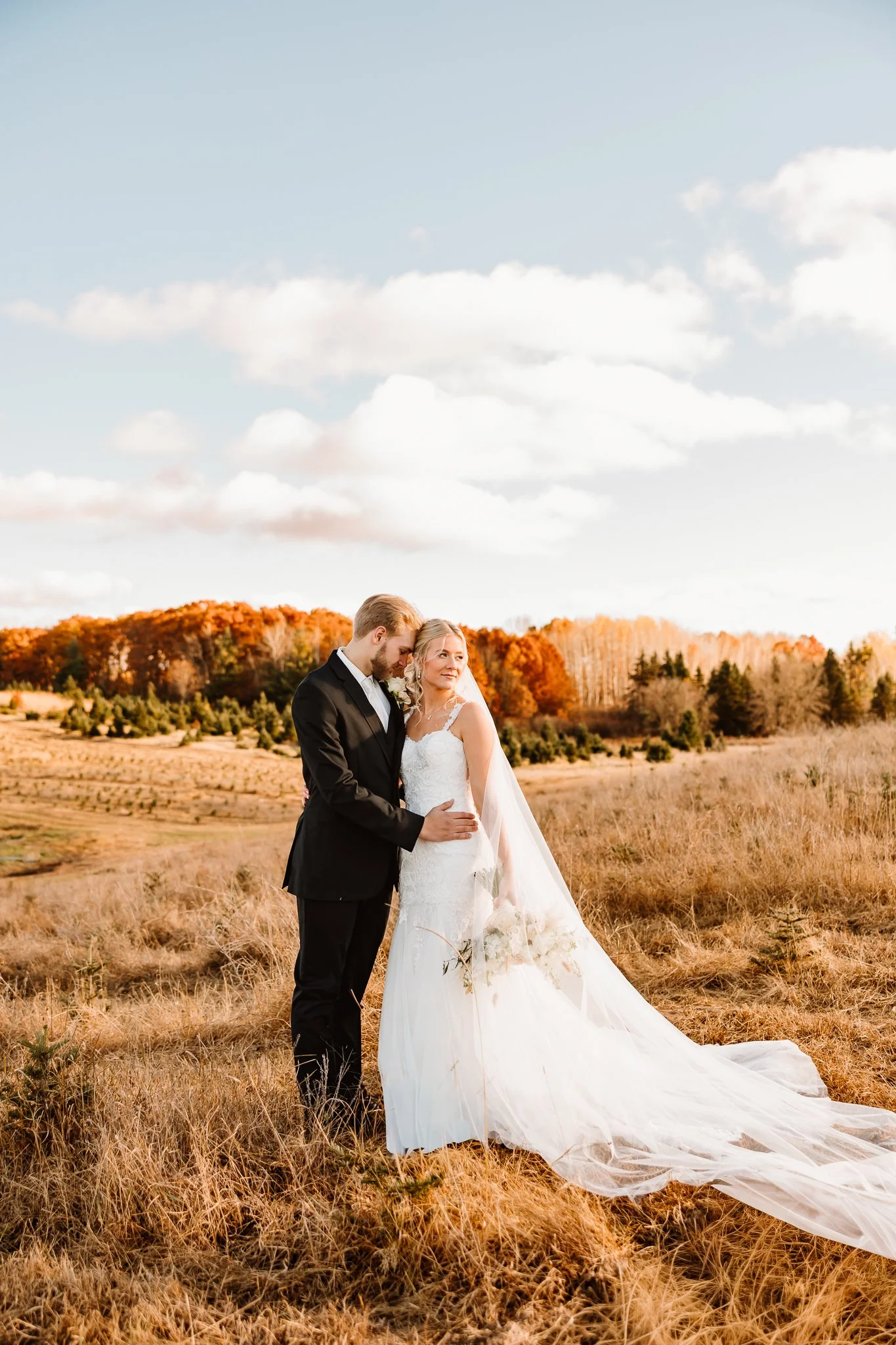 A newlywed couple in wedding attire standing in a field during autumn, with orange trees and a partly cloudy sky in the background.