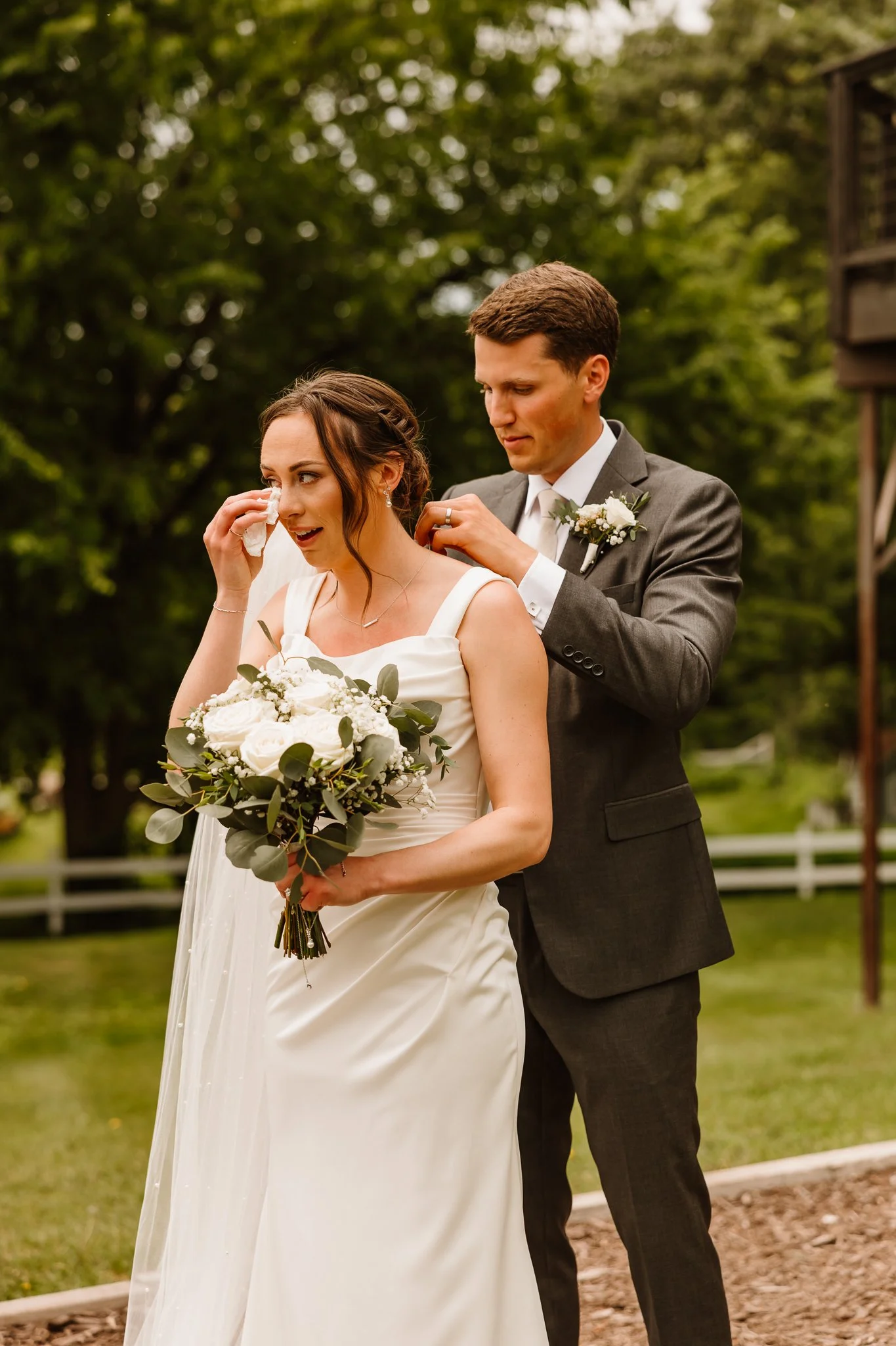A bride and groom sharing an emotional moment outdoors, the groom wiping away tears from the bride's face with a tissue, holding a bouquet of white flowers, during their wedding ceremony.