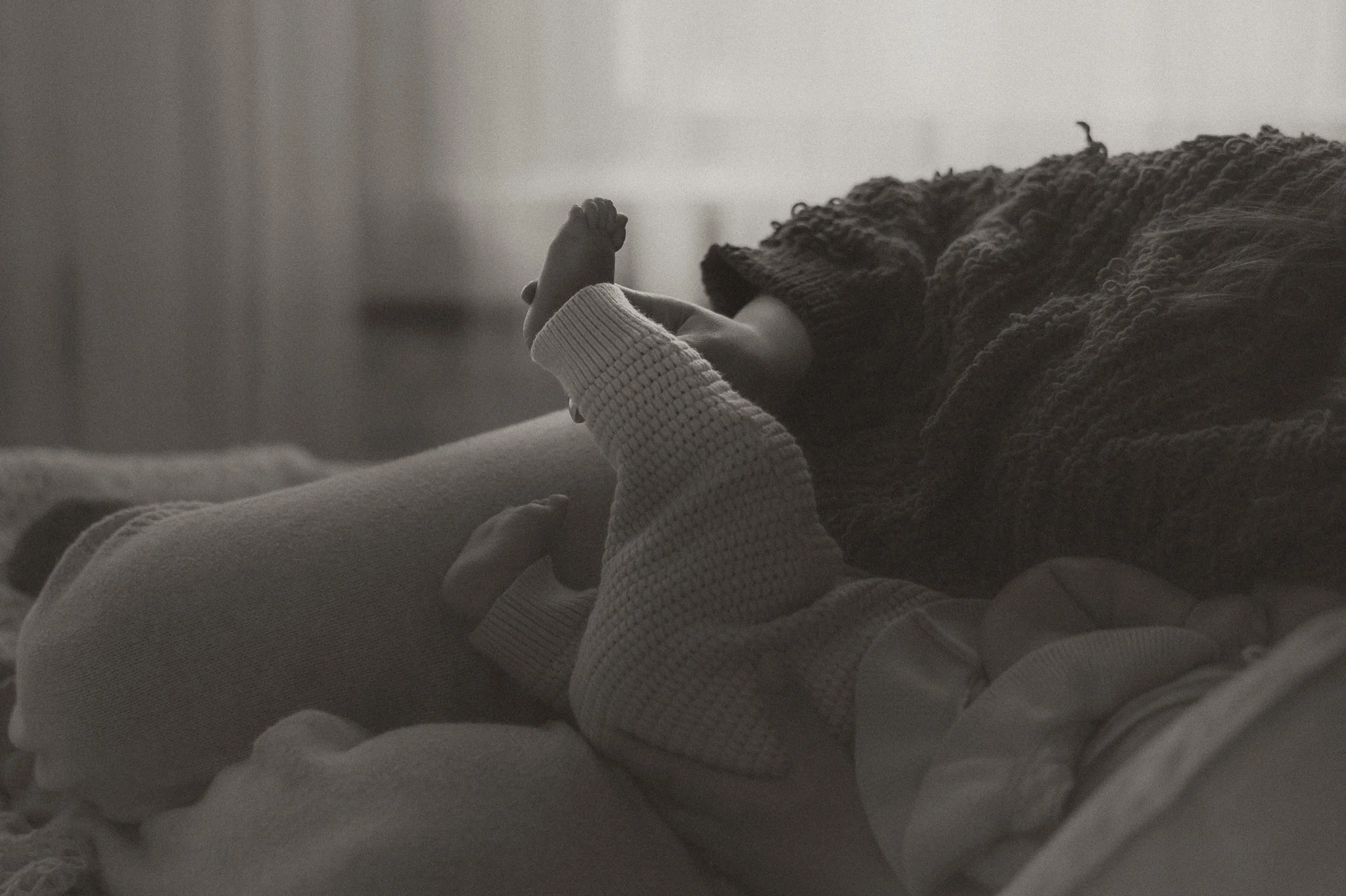 A person is lying on a bed, holding a baby's hand, with their face resting on a pillow. The scene appears calm and intimate, captured in black and white.