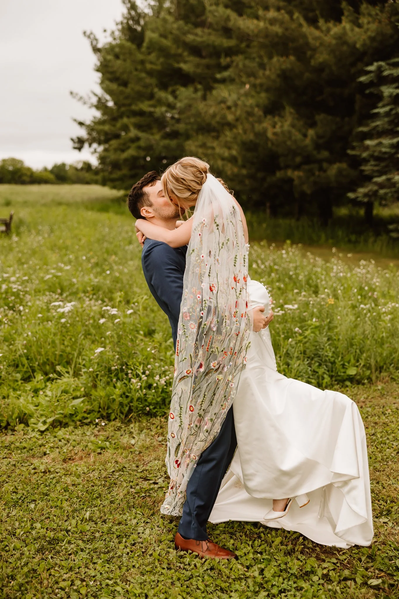 A couple dressed in wedding attire share a romantic moment outdoors. The groom, in a navy suit, holds the bride, who is wearing a white wedding gown and a floral embroidered veil. The bride has her arms around the groom's neck, and they are about to 