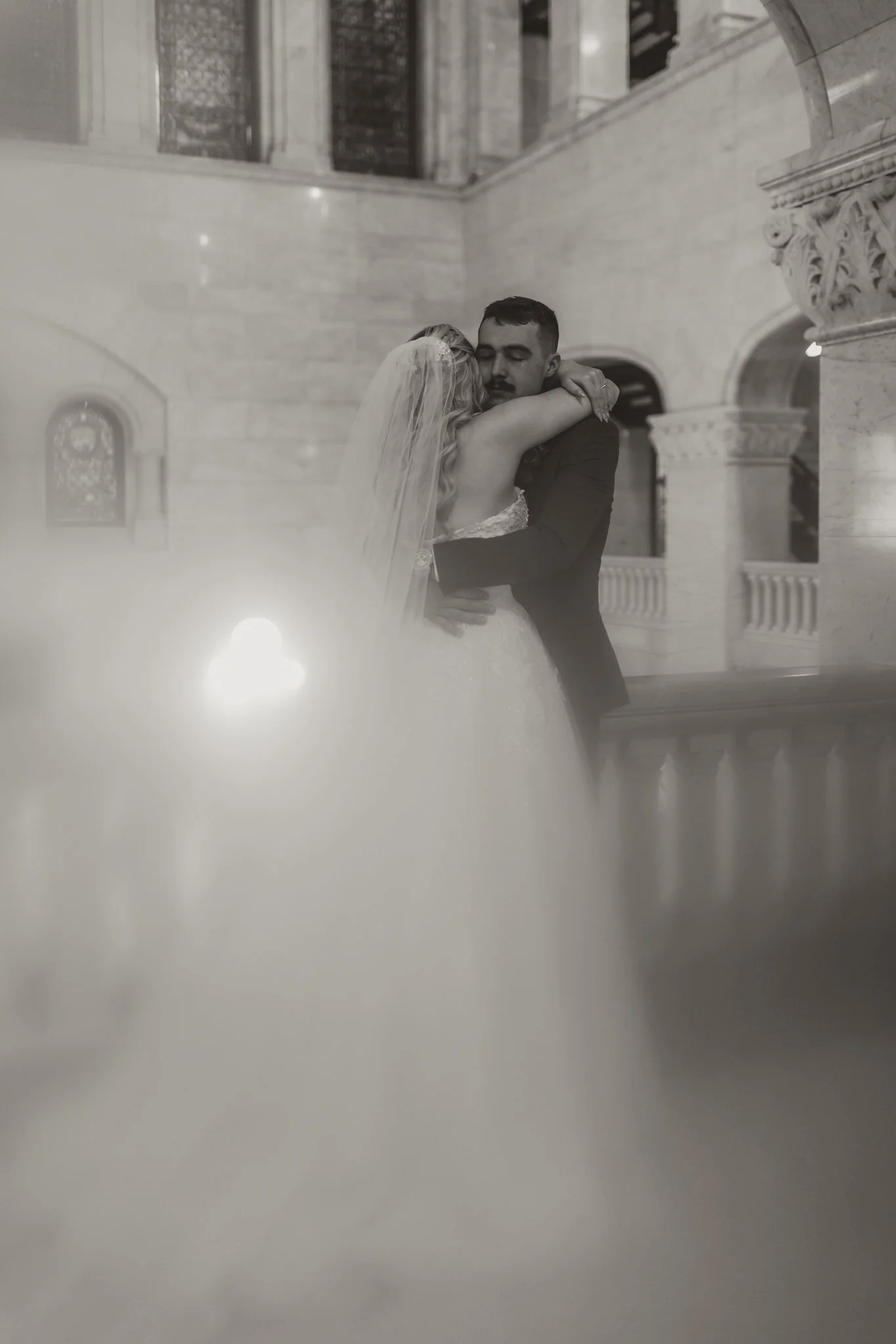 A bride and groom embrace in a stairwell of a grand building, with stone architecture and stained glass windows, in black and white.