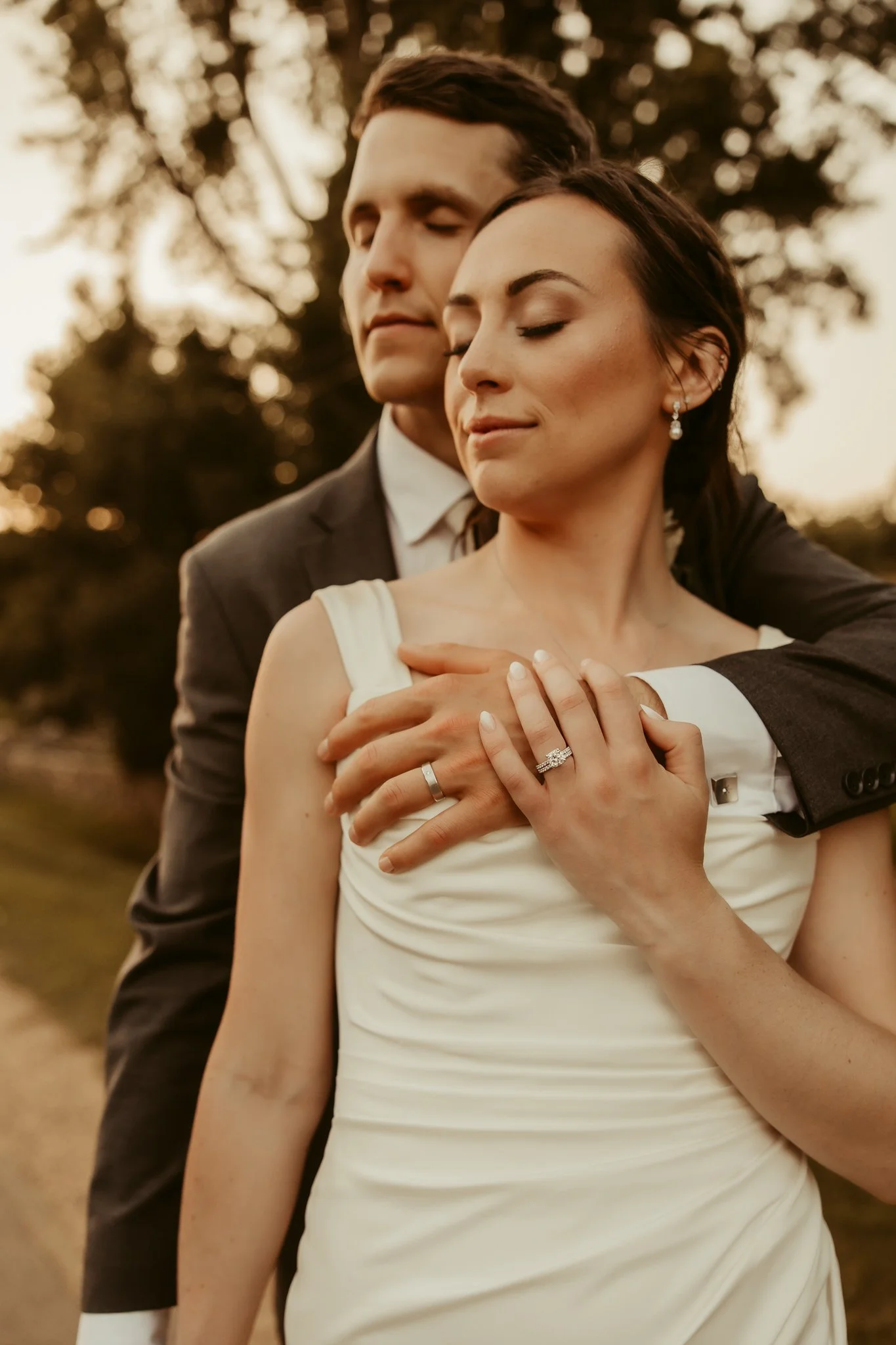 A bride and groom with eyes closed embracing outdoors at sunset, showing their wedding rings.