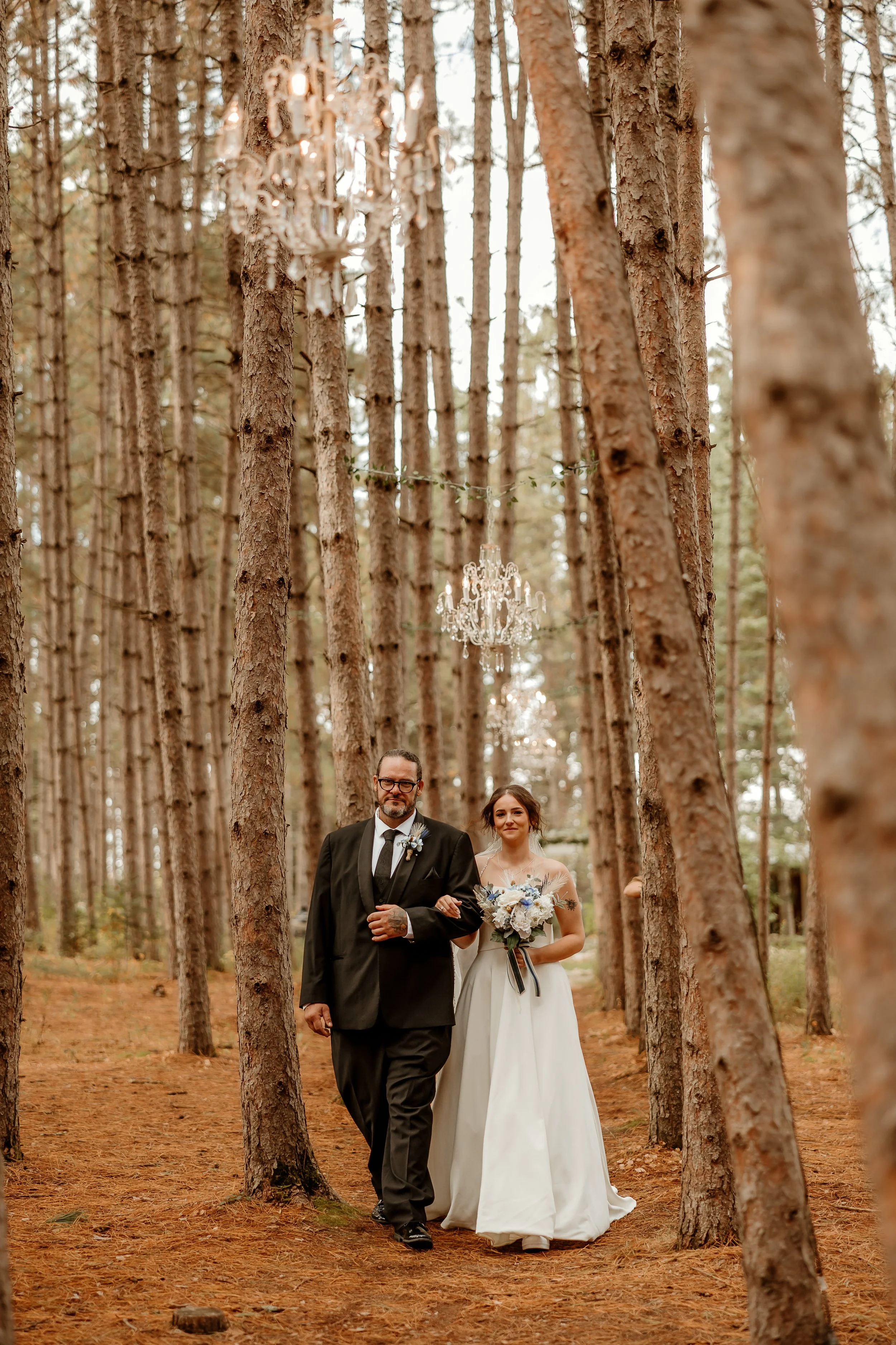 A bride in a white wedding dress walking down a forest path with a man in a black tuxedo. The bride holds a bouquet of white and blue flowers. Crystal chandeliers are hanging from the trees above.