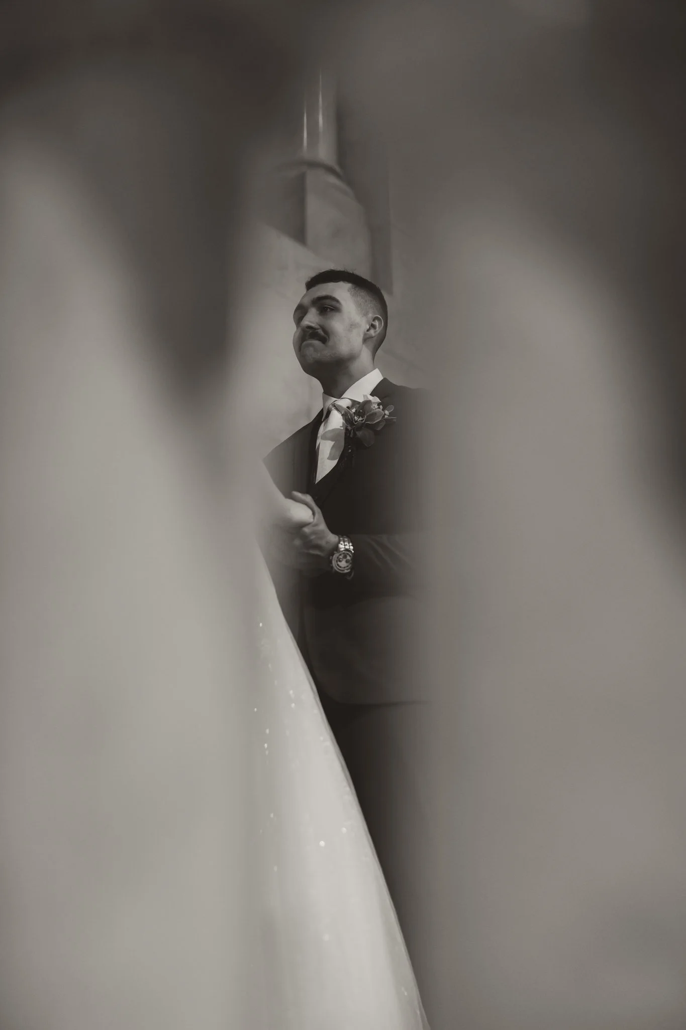 A black and white photo of a man in a tuxedo with a boutonnière, holding his chest, seen through a narrow opening around two blurred figures, likely a bride and another person, in a church or similar setting.