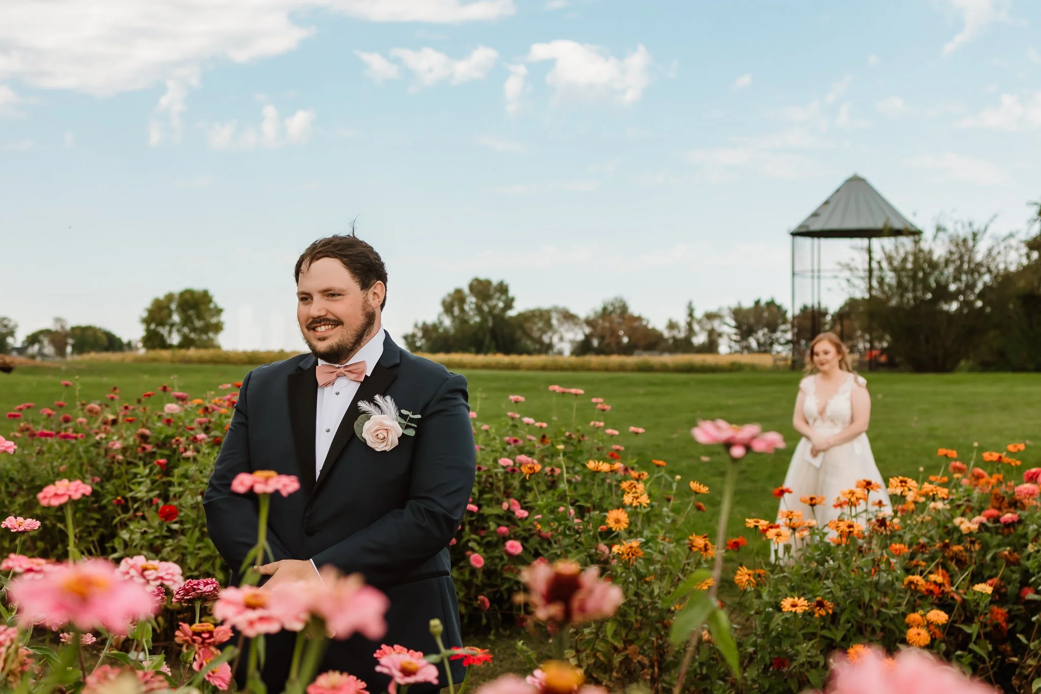 A groom in a black tuxedo with a pink bow tie and boutonniere, smiling and standing among pink and orange flowers outdoors. A bride in a white wedding dress is in the background, standing on green grass with a pavilion and trees visible under a partl