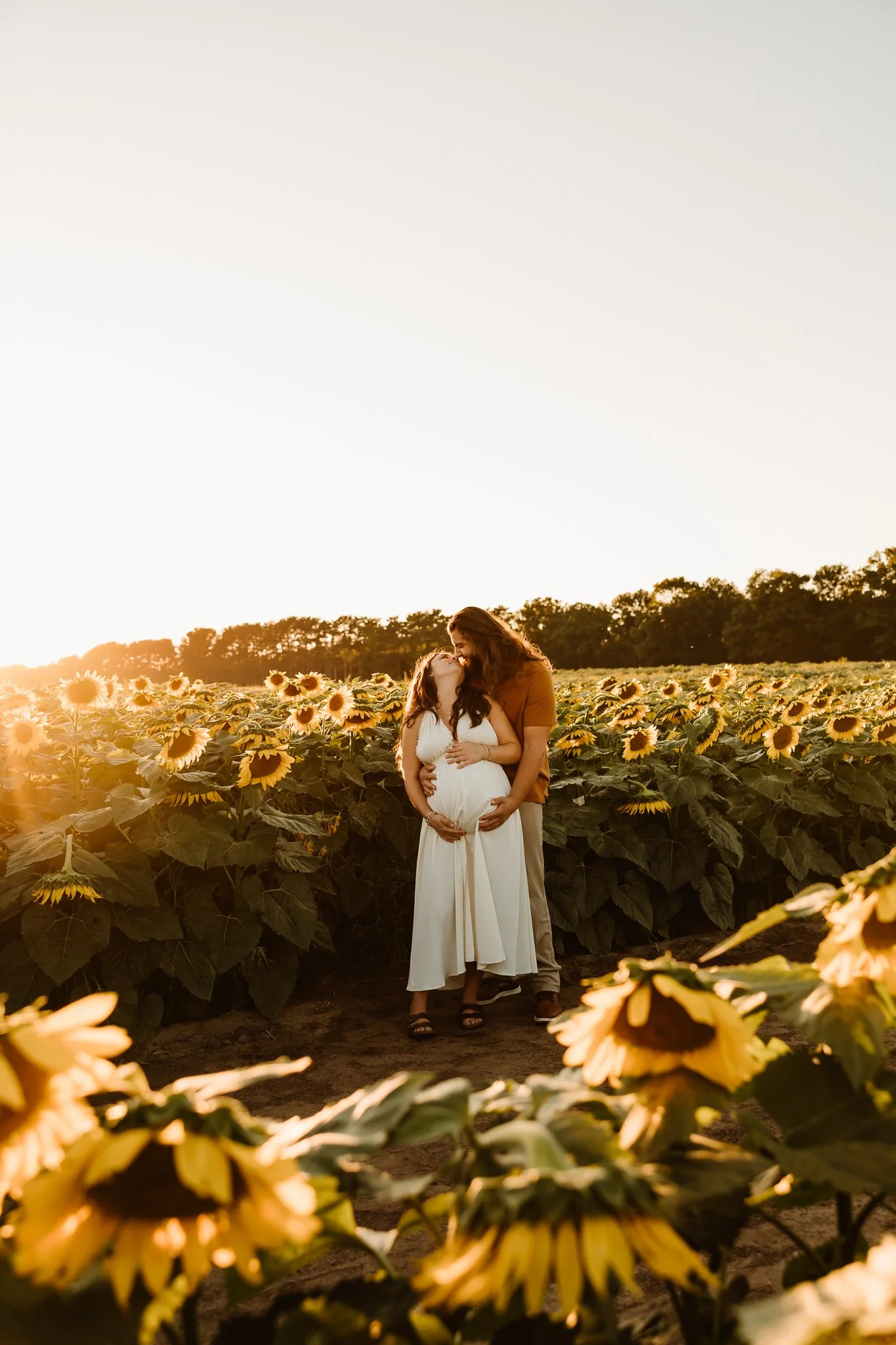A pregnant woman and a man standing in a sunflower field at sunset, sharing a tender moment with the woman holding her belly and the man embracing her from behind.