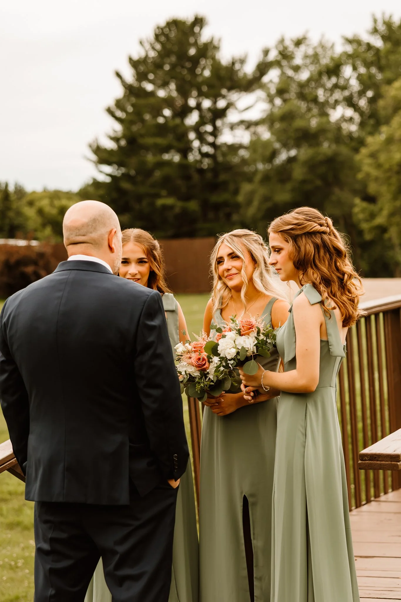 A bride and two bridesmaids in matching sage dresses holding bouquets, standing outside and talking to a groom in a black suit during a wedding ceremony.