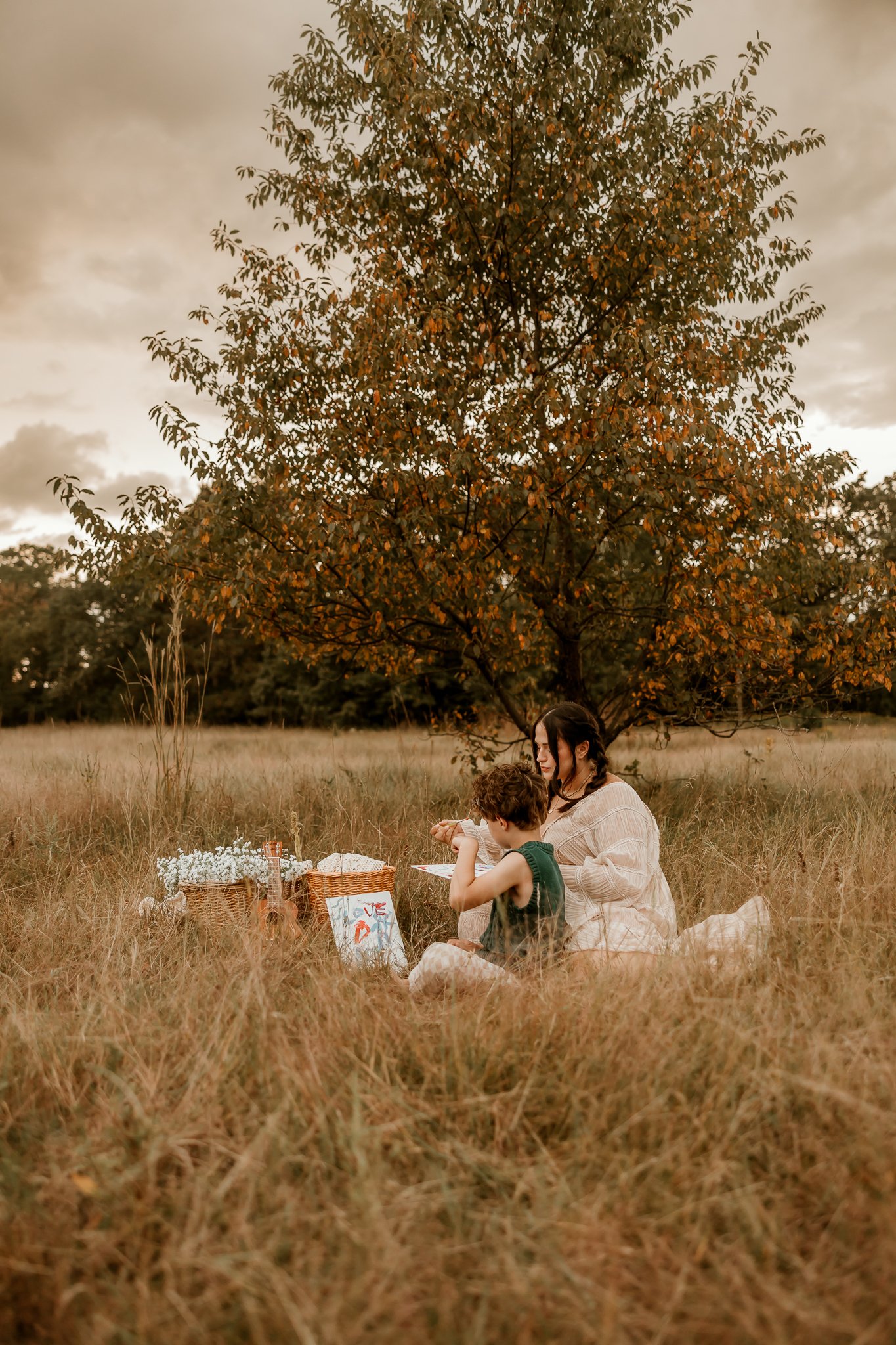 A woman and a young child sitting on a blanket in a grassy field under a tree, having a picnic with baskets and flowers, during a cloudy day.
