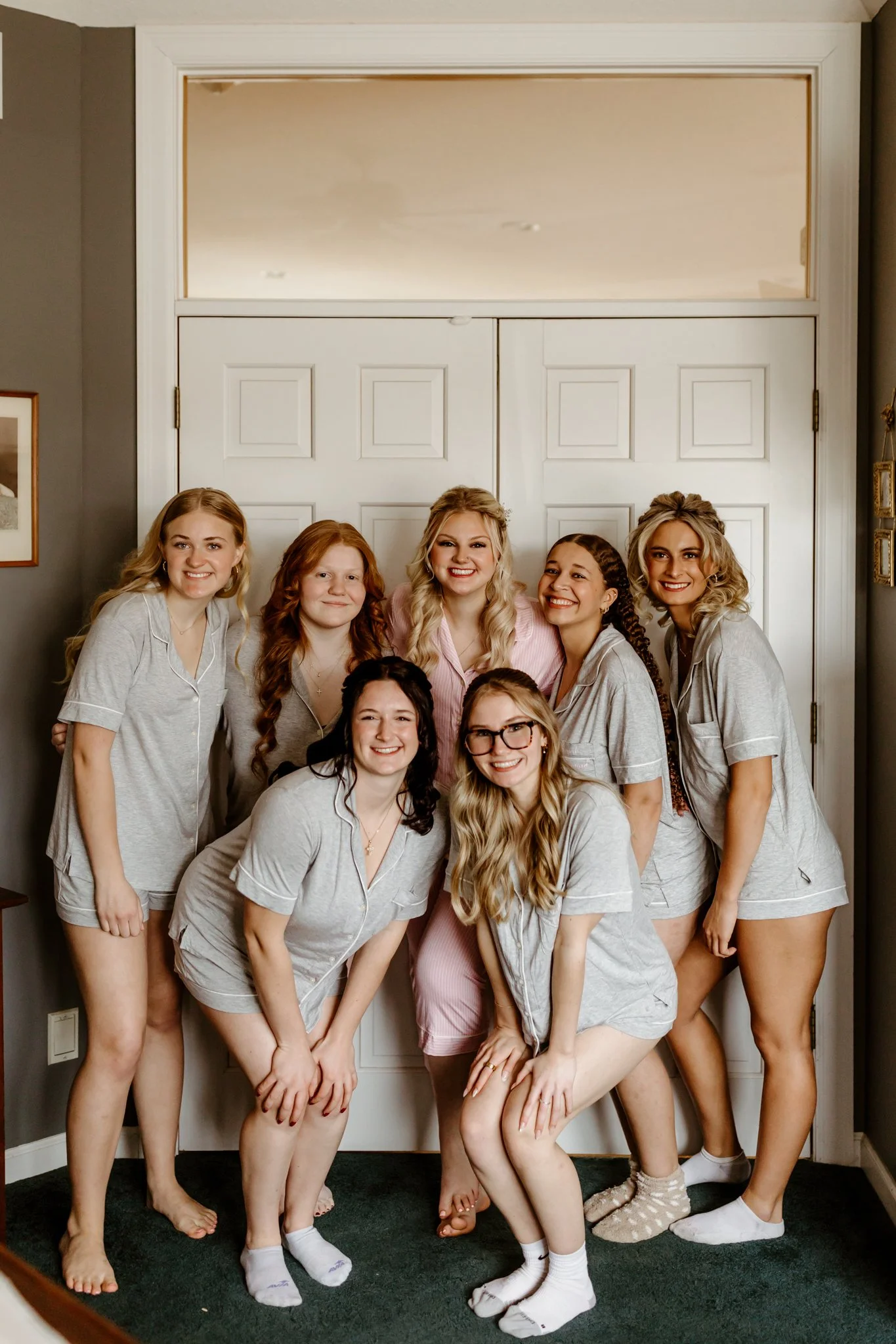 Group of seven women in pajamas smiling inside a home, standing in front of a white paneled door.