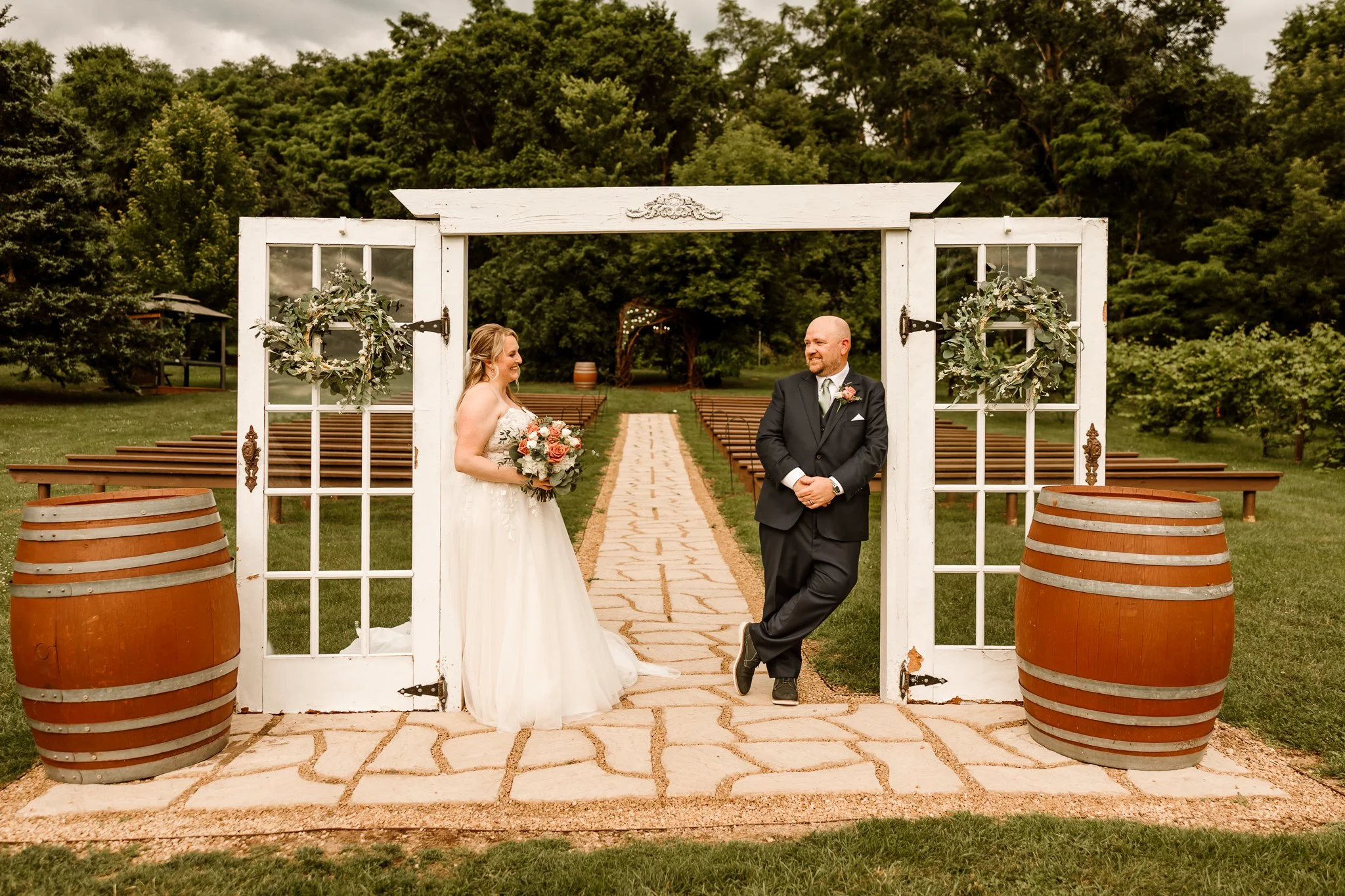 Bride and groom standing at an outdoor wedding ceremony, smiling at each other, framed by a white decorative arch with green wreaths, set on a stone pathway in a lush green gardenHope Glen Farm Cottage Grove Minnesota 