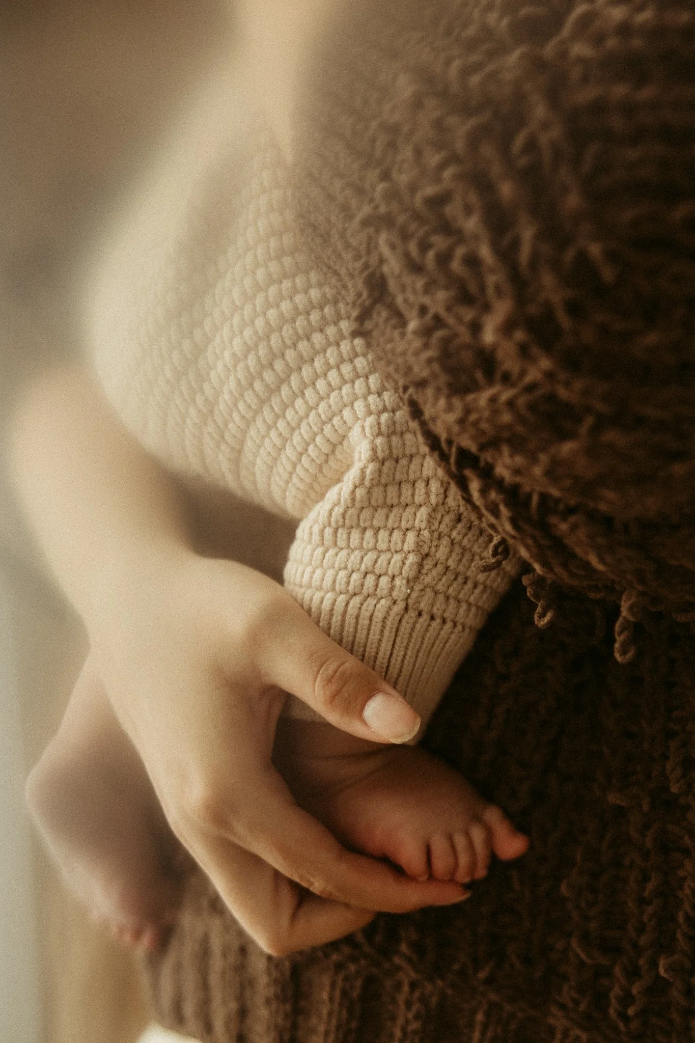 A close-up of a baby's hand gripping an adult's finger, with a textured beige sleeve and brown fuzzy blanket or sweater nearby.