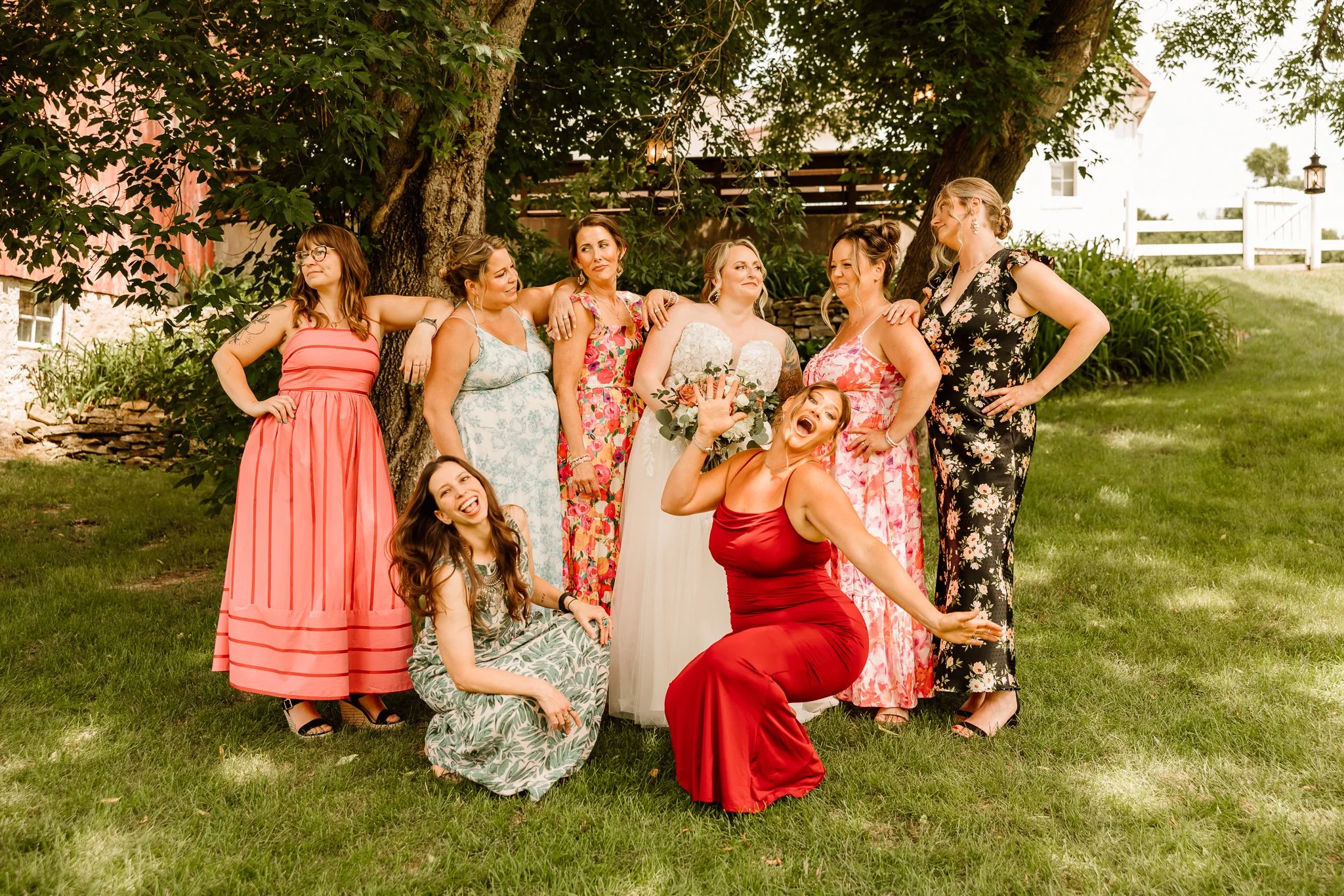 Group of women and a girl in colorful dresses posing outdoors under a large tree, celebrating a wedding, with one woman in a white wedding dress in the center holding a bouquet.