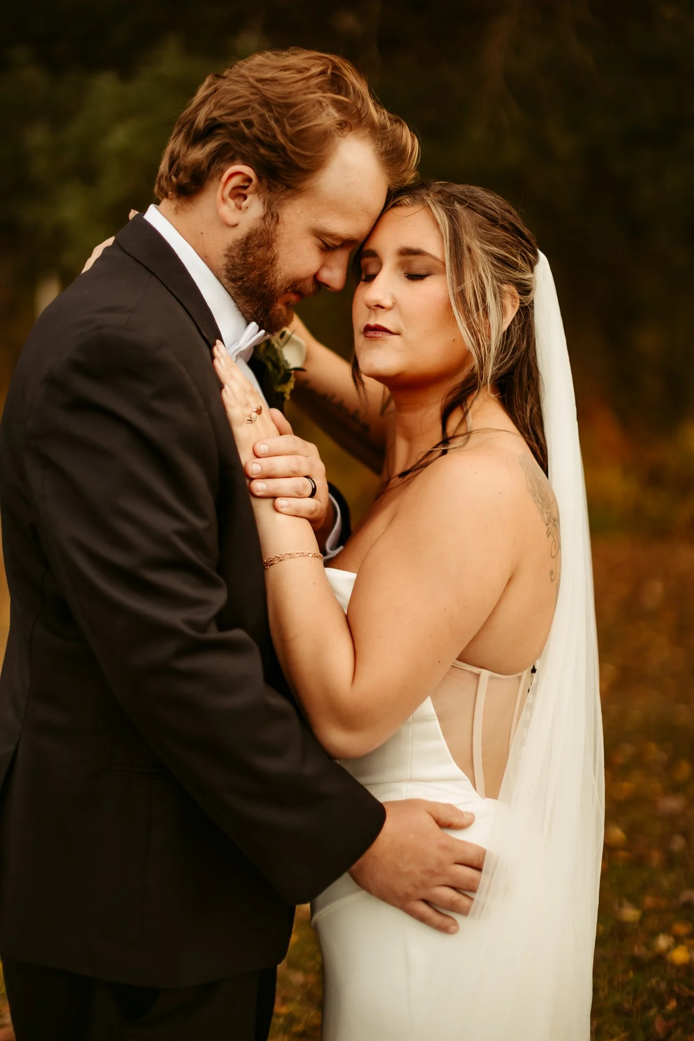 A couple dressed in wedding attire, with their foreheads and noses touching, standing close with their eyes closed, outdoors during autumn.