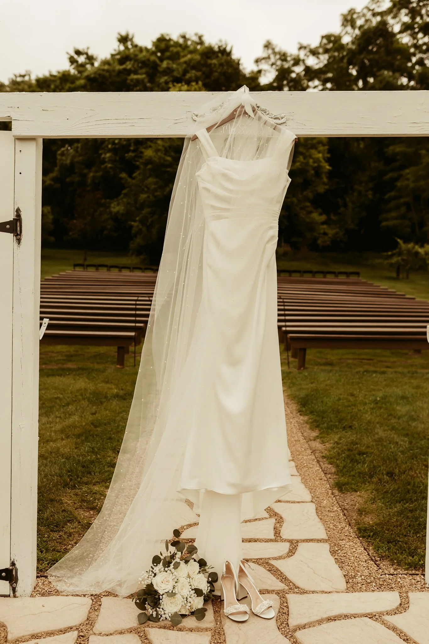 Wedding dress hanging on a wooden gate with a veil, bouquet of flowers, and pair of heels placed on the ground in front of the dress, outdoors with green trees in the background.