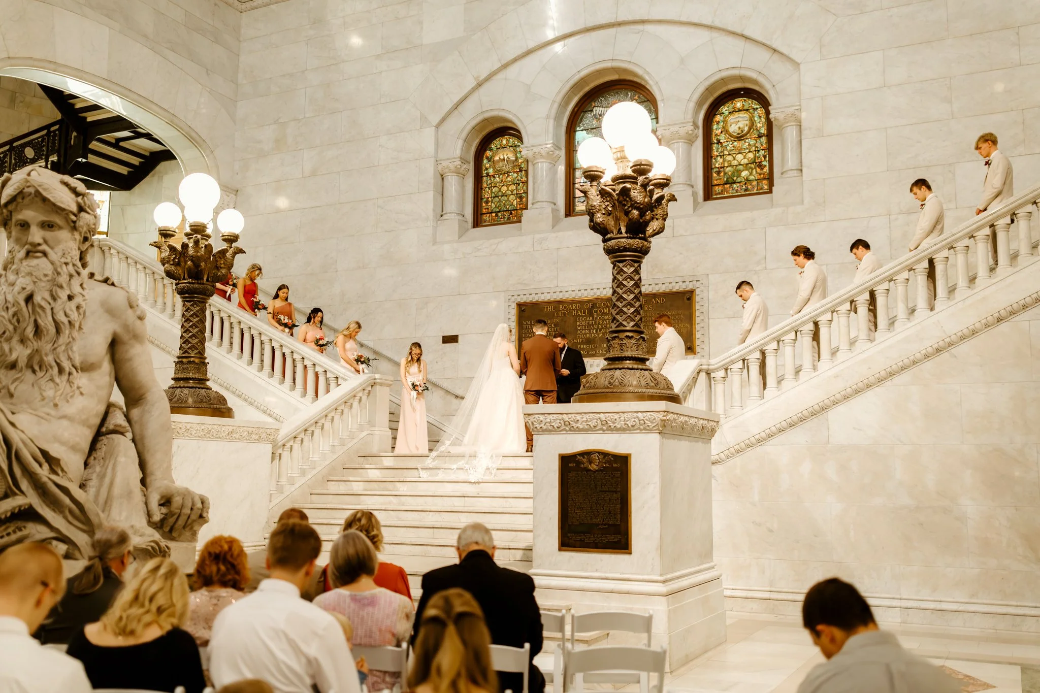A wedding ceremony taking place inside a grand marble hall with a staircase, stained glass windows, and ornate decor. The bride and groom stand before an officiant, surrounded by bridesmaids and groomsmen.