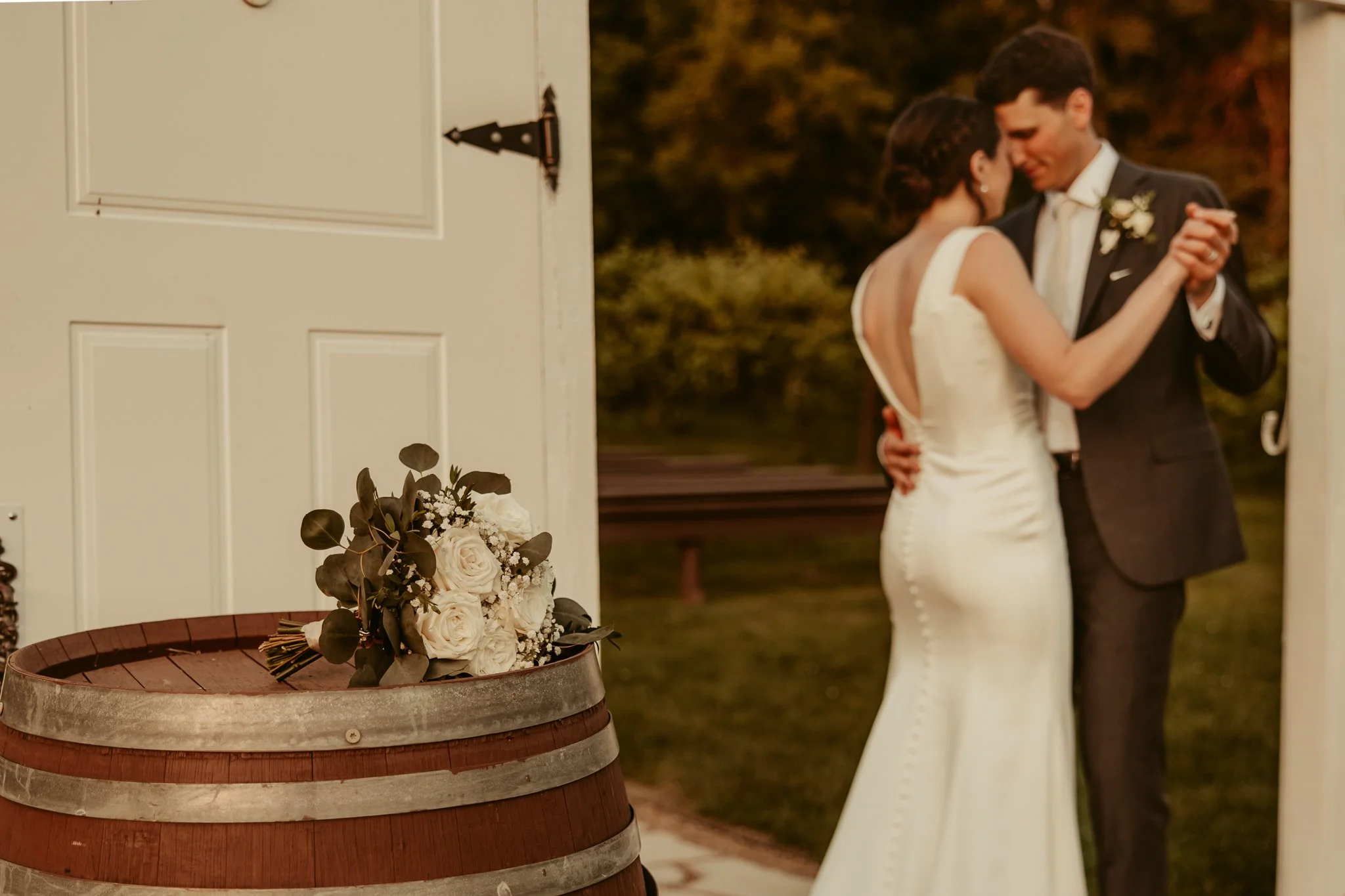 Wedding bouquet resting on a wooden barrel with a bride and groom dancing in the background.