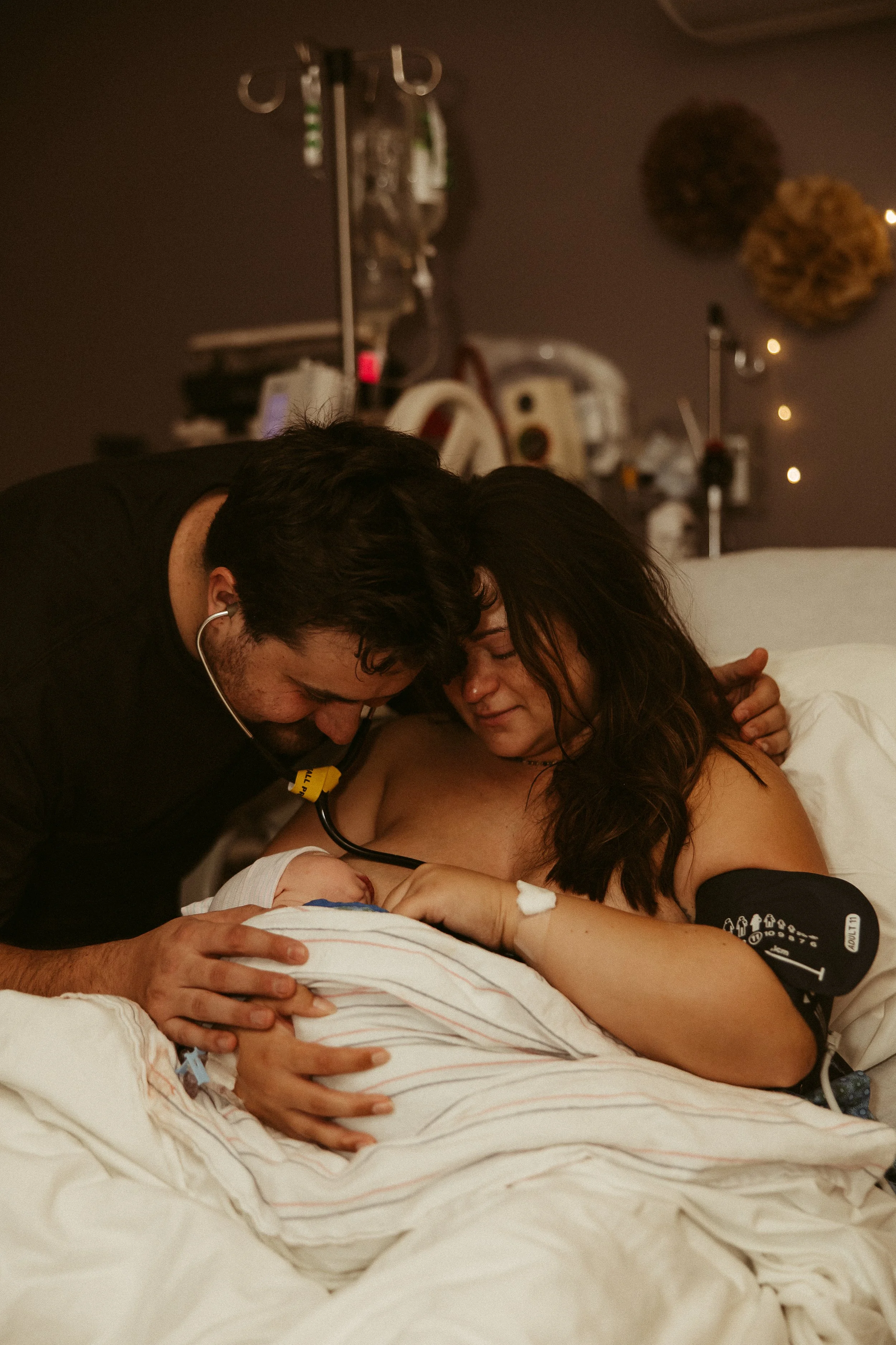 A man and a woman, both appearing distressed and emotional, are in a hospital room with a newborn baby. The woman is lying in a hospital bed with medical monitoring devices attached. The man is leaning over her, holding her hand, with a stethoscope a