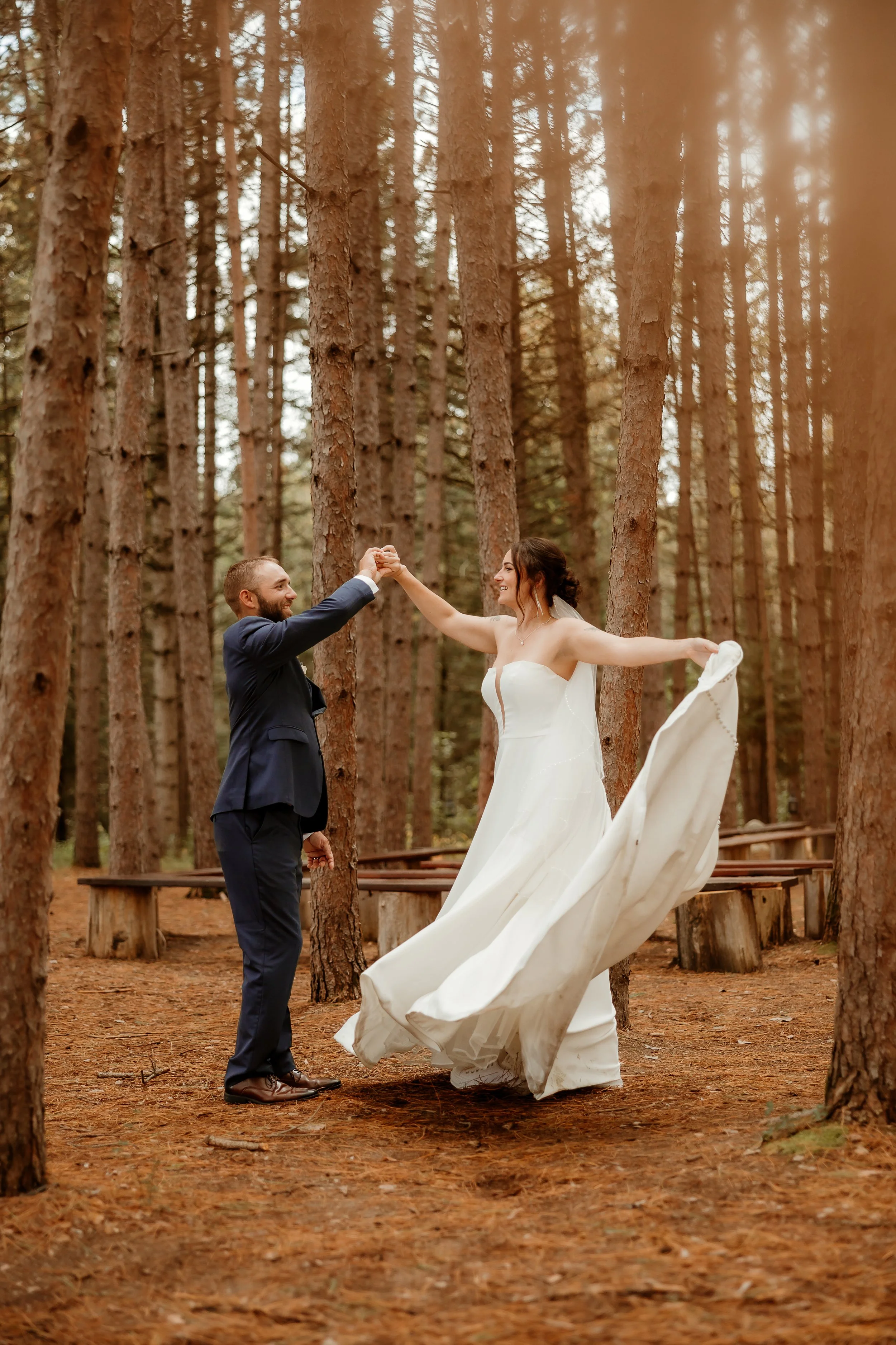 A bride and groom dancing in a wooded forest, with the bride wearing a white wedding dress and the groom in a dark suit.