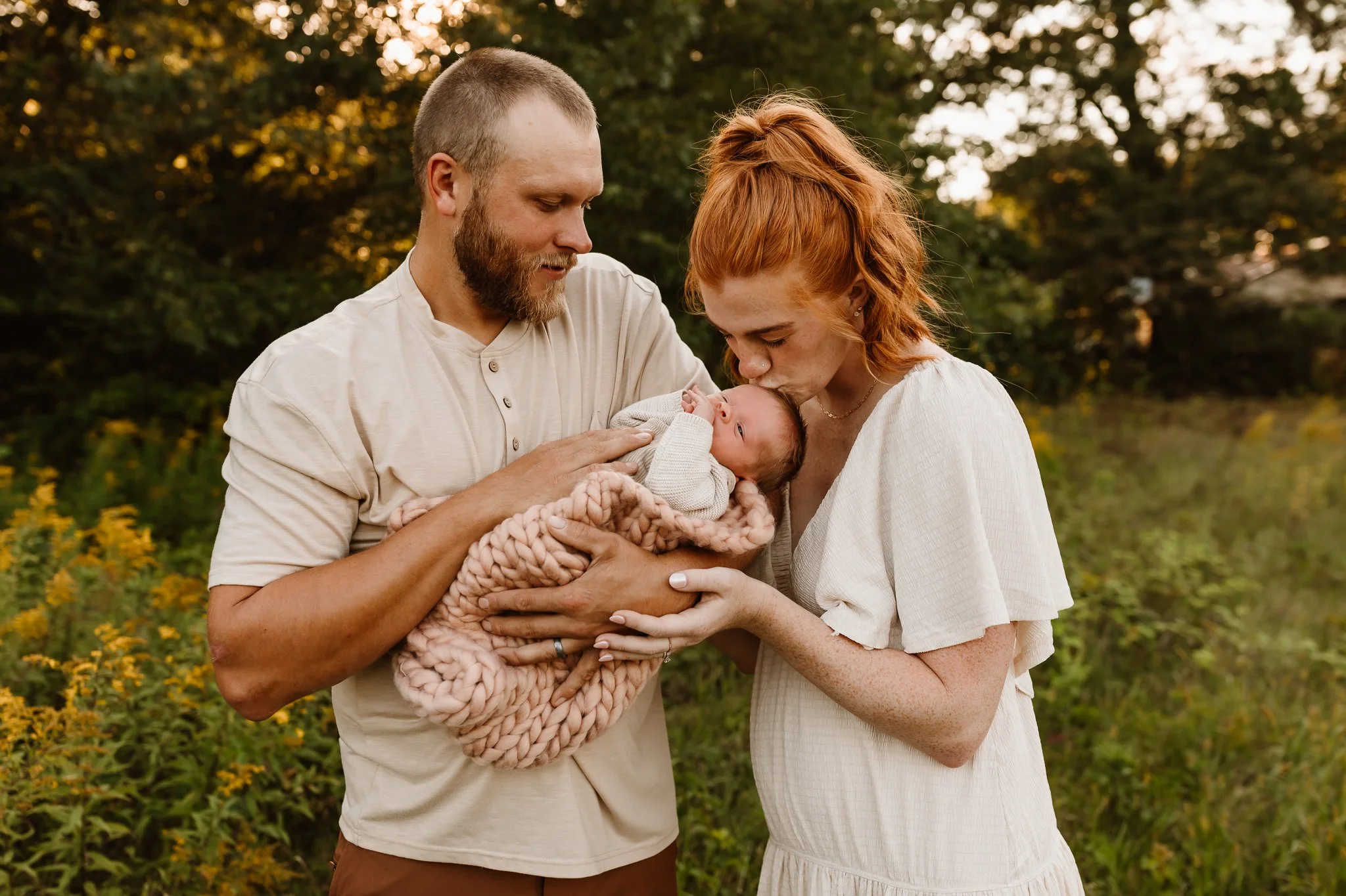 A man and a woman hold a newborn baby outdoors during sunset, with trees and yellow flowers in the background.